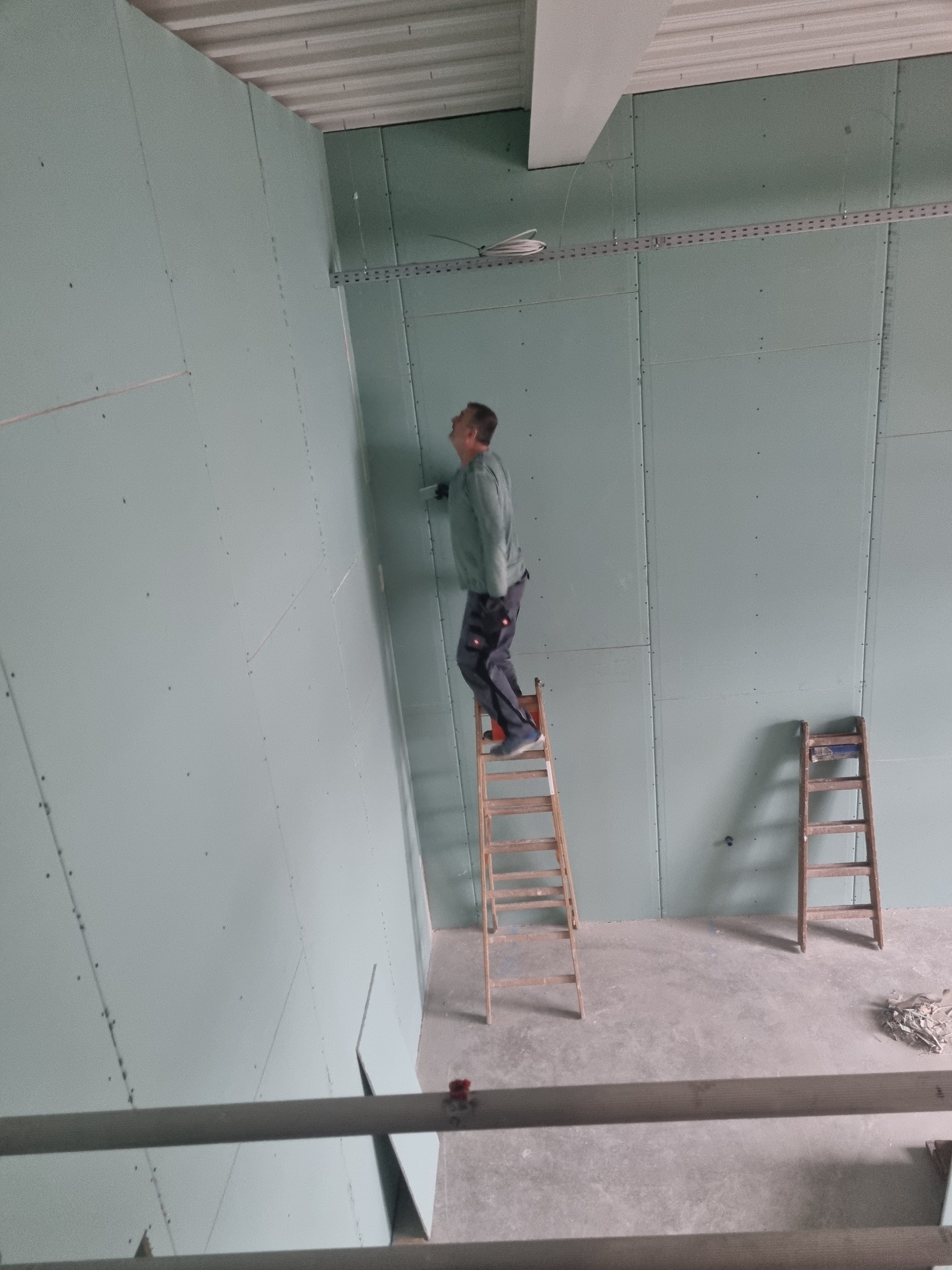 Pracownik na drabinie montuje płyty karton gips na ścianie, widoczne ślady po wkrętach, druga drabina oparta o ścianę w tle.