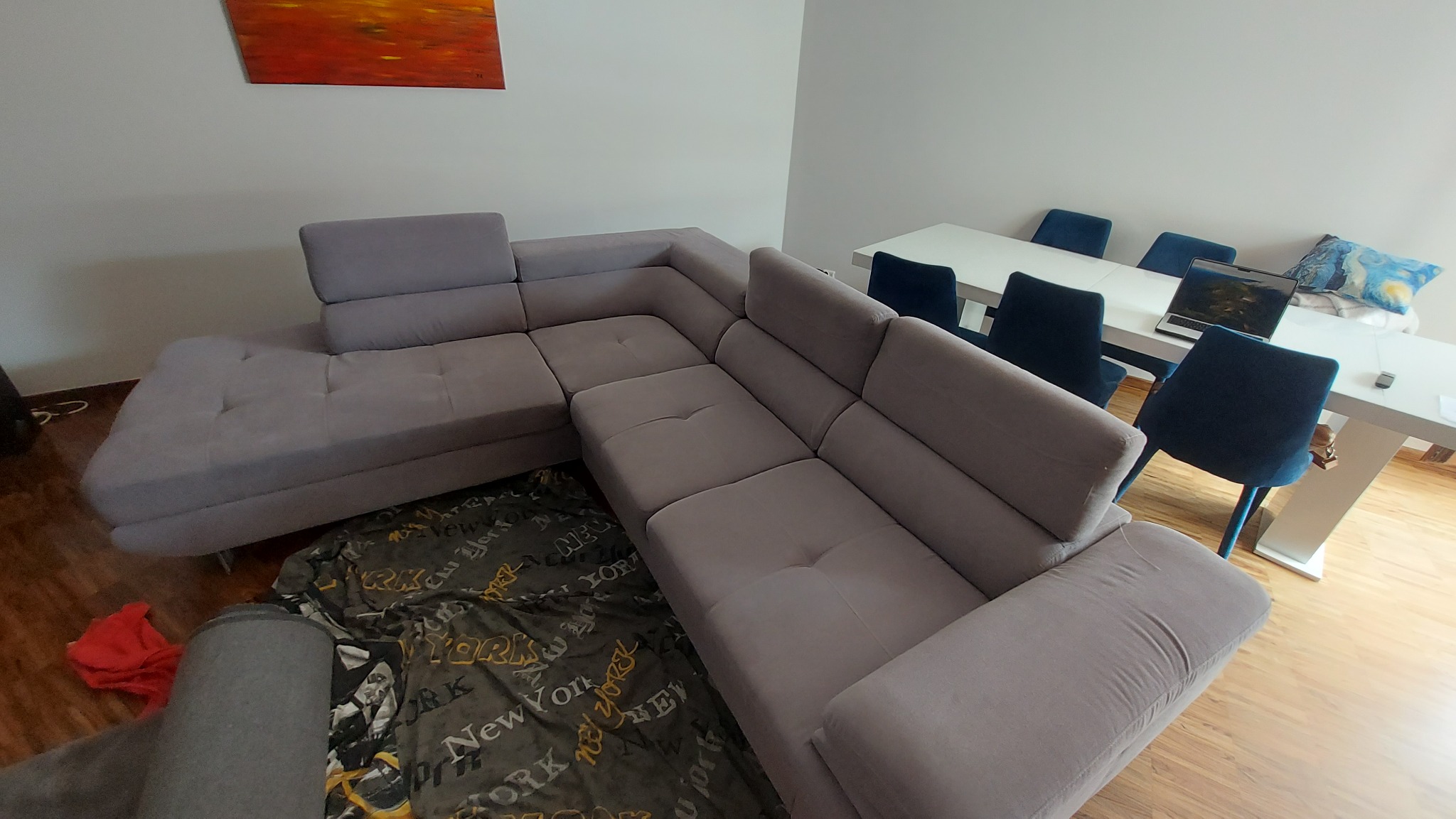 Szary narożnik z tapicerką w salonie, na podłodze koc z napisem New York, w tle stół z krzesłami i laptopem.