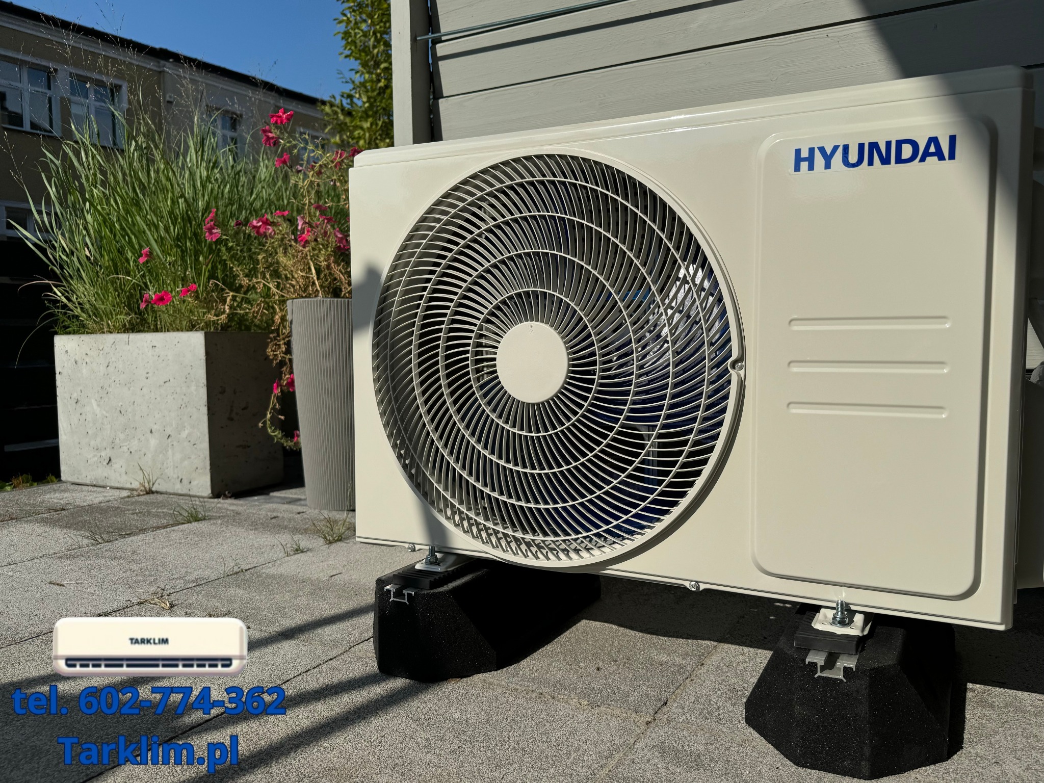 Zewnętrzna jednostka klimatyzacji Hyundai zamontowana na betonowym patio, z widocznymi donicami z zielenią w tle.