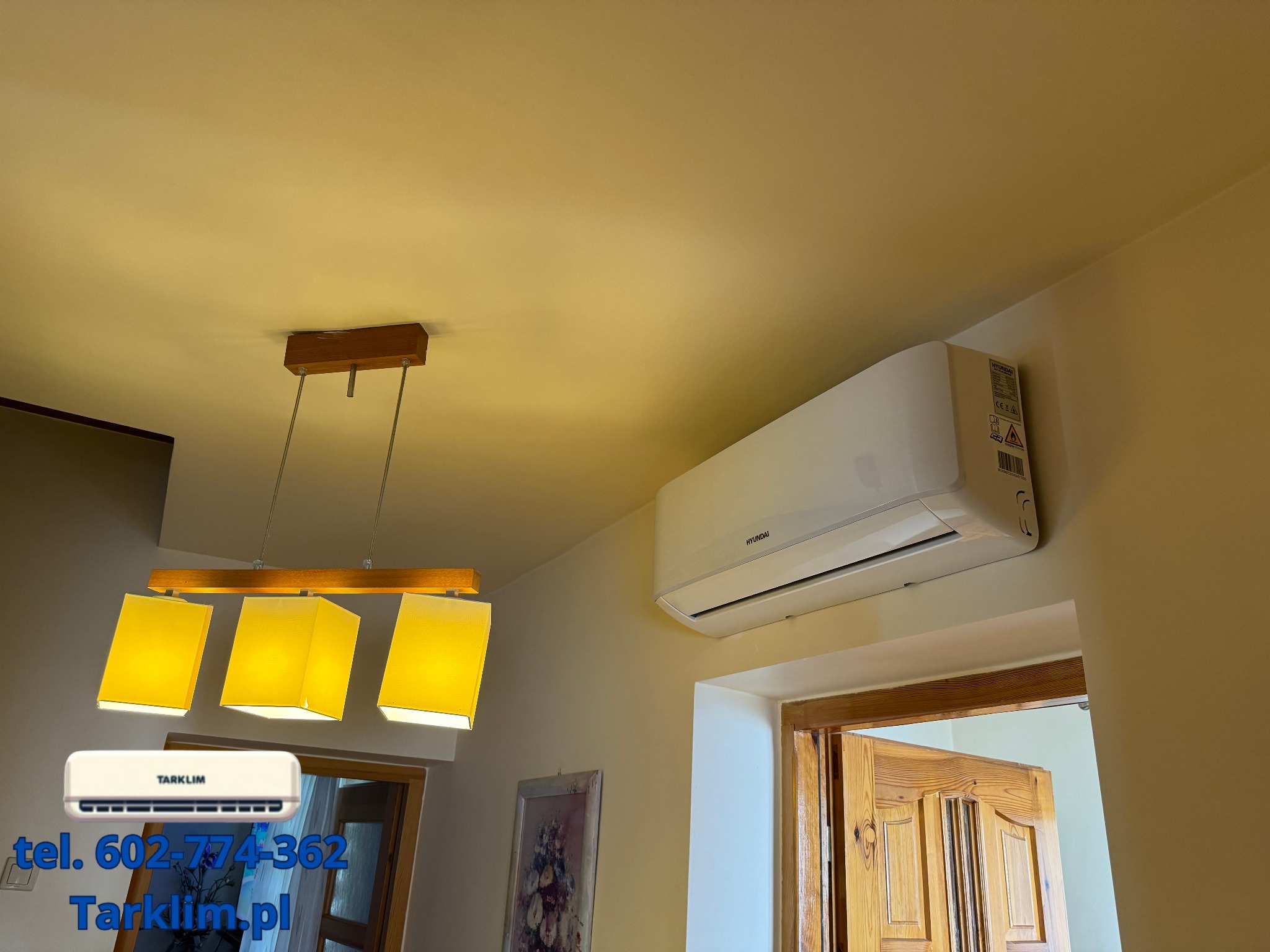 Zamontowana jednostka klimatyzacji Hyundai w jasnym wnętrzu, widoczny drewniany żyrandol z trzema kloszami i fragment drewnianych drzwi.