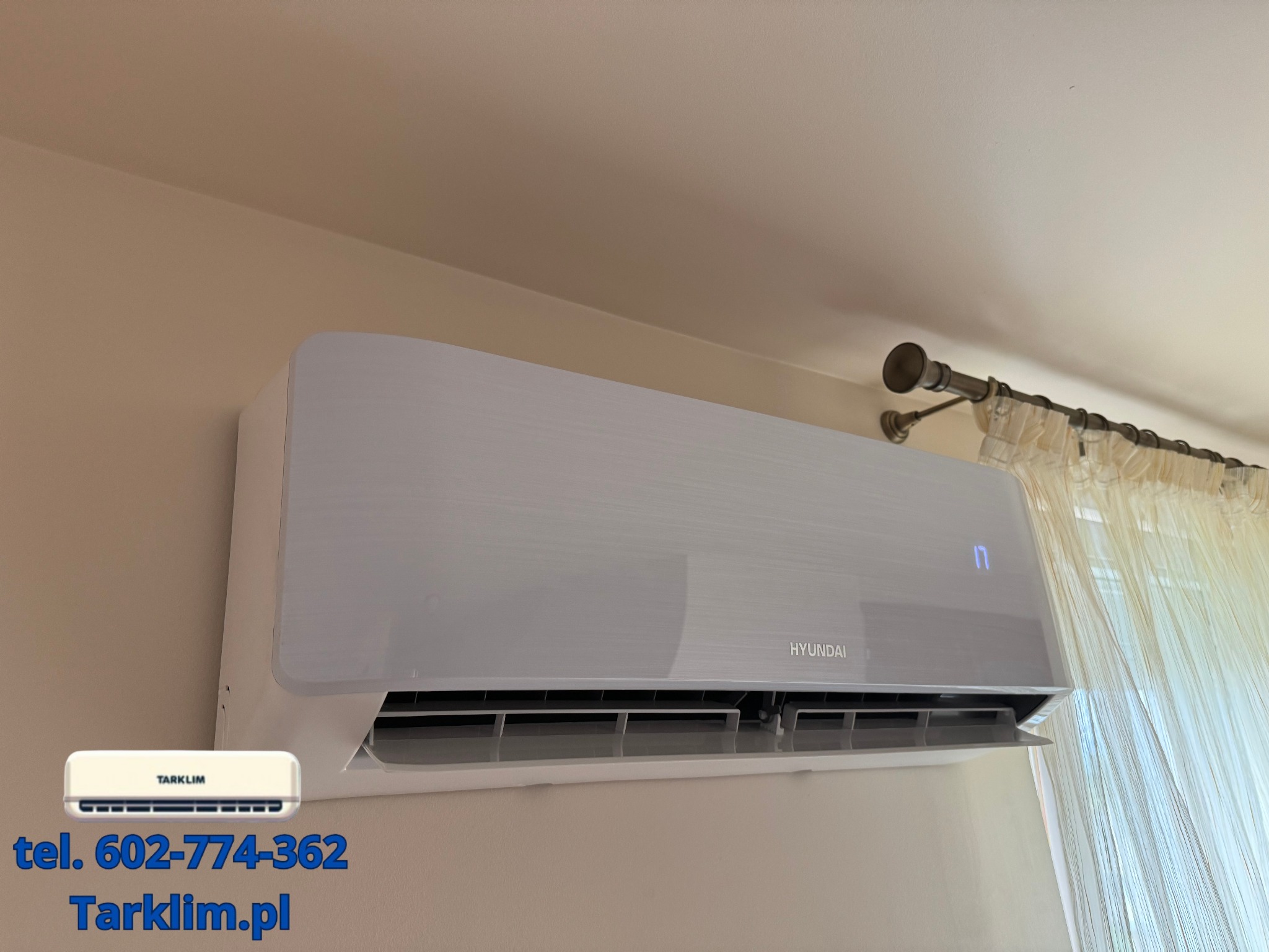 Biała jednostka klimatyzacji Hyundai zamontowana na ścianie w pomieszczeniu z zasłonami, widoczne logo firmy TARKLiM z numerem telefonu i adresem strony internetowej w lewym dolnym rogu.