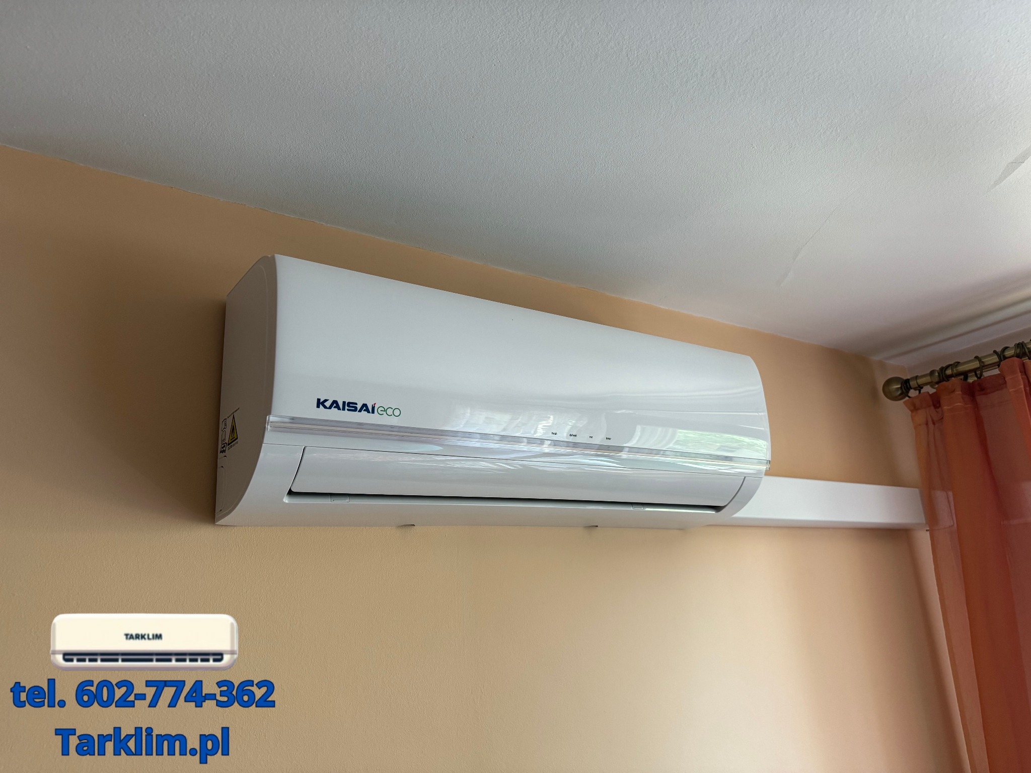 Biały klimatyzator Kaisaieco zamontowany na beżowej ścianie w pomieszczeniu z zasłoniętym oknem.