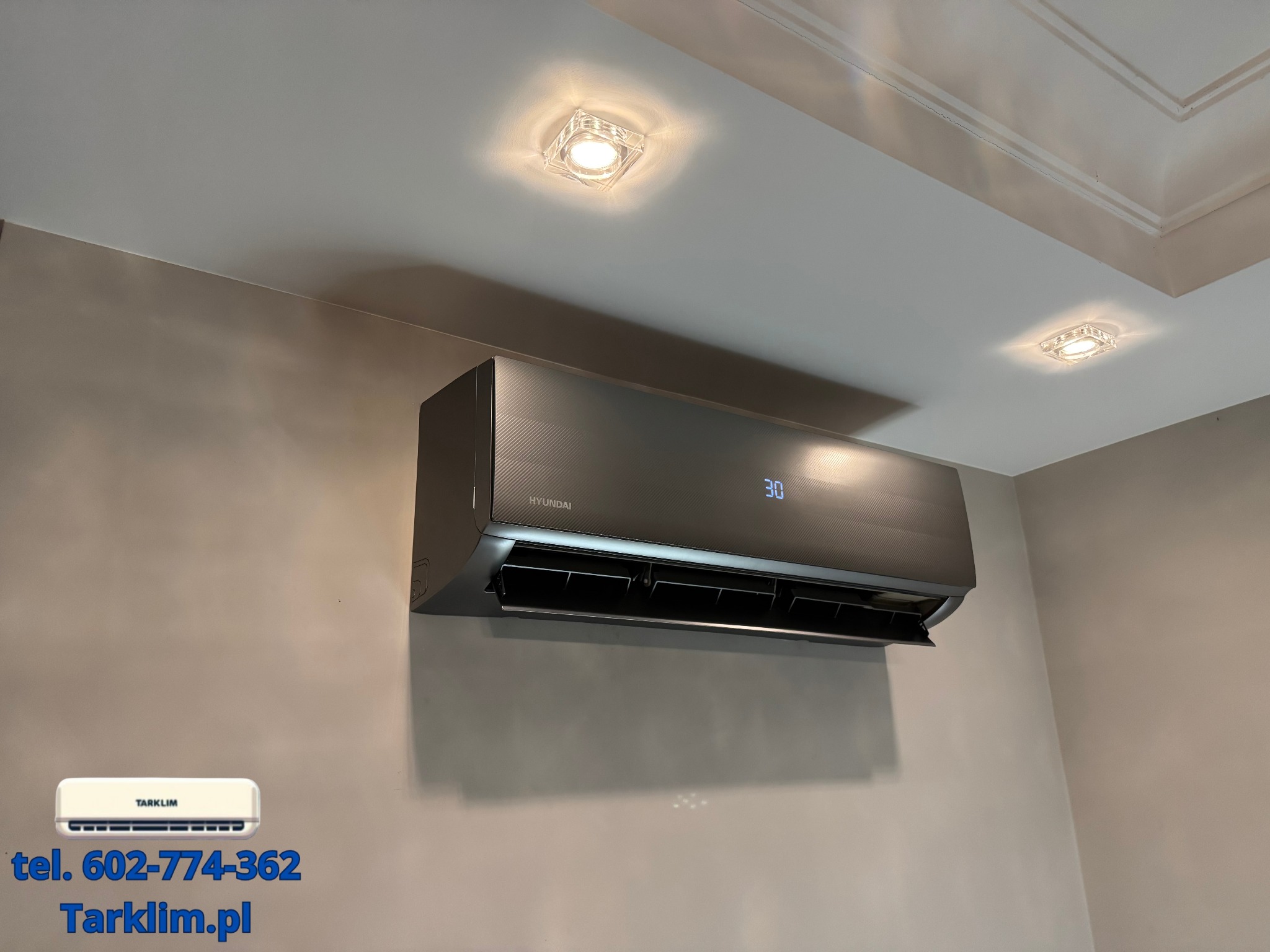 Zamontowany na ścianie klimatyzator Hyundai w kolorze grafitowym, z wyświetlaczem pokazującym temperaturę 30 stopni, na tle beżowej ściany i sufitu z kwadratowymi lampami.