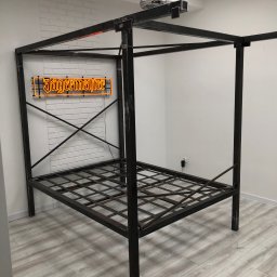 Daniel Leigna - Metalowa rama łóżka z baldachimem w surowym, industrialnym stylu, z neonem Jägermeister na tle ściany z białej cegły.