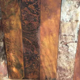 Rdzewiejące, pionowe metalowe panele z wyraźną teksturą i zróżnicowaną kolorystyką odcieni brązu, pomarańczy i szarości, ukazujące proces korozji