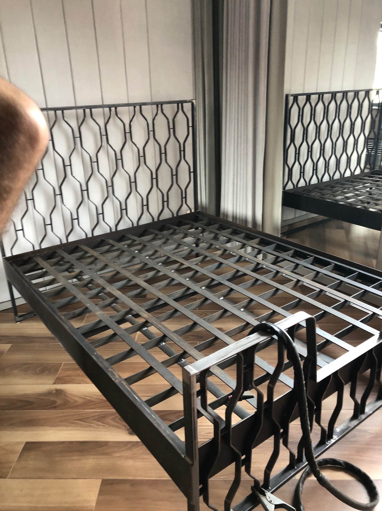 Metalowa rama łóżka z ozdobnym zagłówkiem i podnóżkiem, spawana konstrukcja kratowa, widoczne ślady spawania, drewnopodobna podłoga w tle.