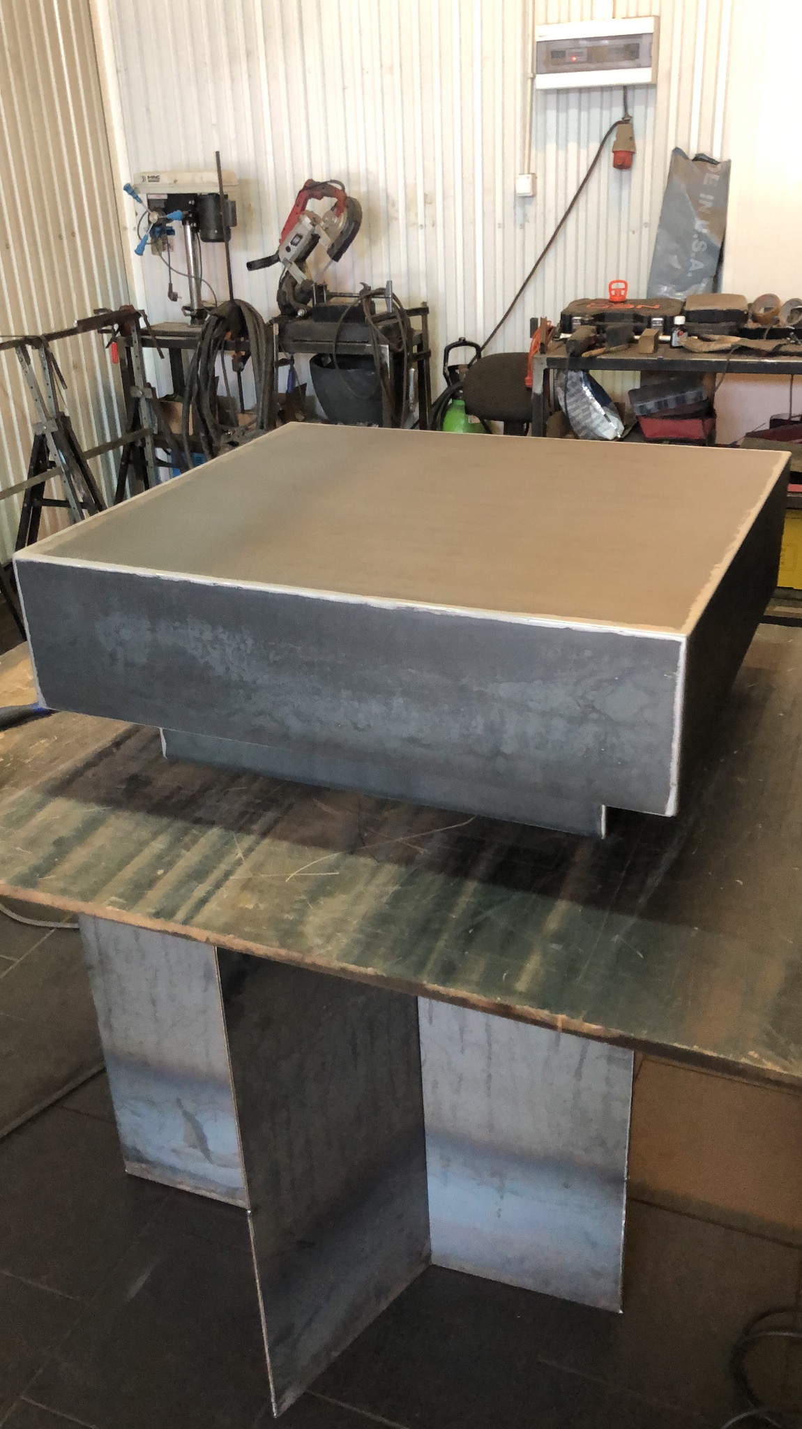 Surowa, spawana konstrukcja ze stali, prawdopodobnie podstawa stołu lub element dekoracyjny, na tle warsztatu z widocznymi narzędziami i urządzeniami.