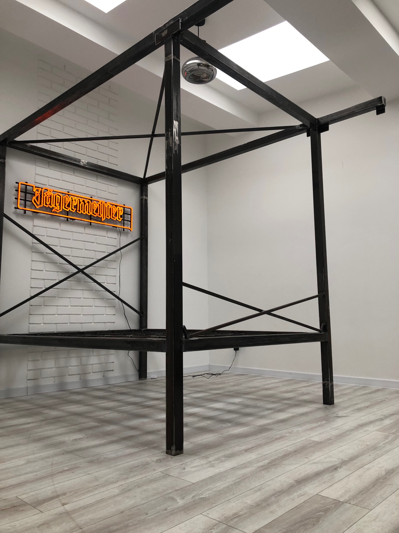 Surowa, metalowa konstrukcja sześcianu, prawdopodobnie pergola lub element dekoracyjny, z widocznymi spawami, umieszczona w minimalistycznym wnętrzu z neonem 'Jägermeister' w tle.