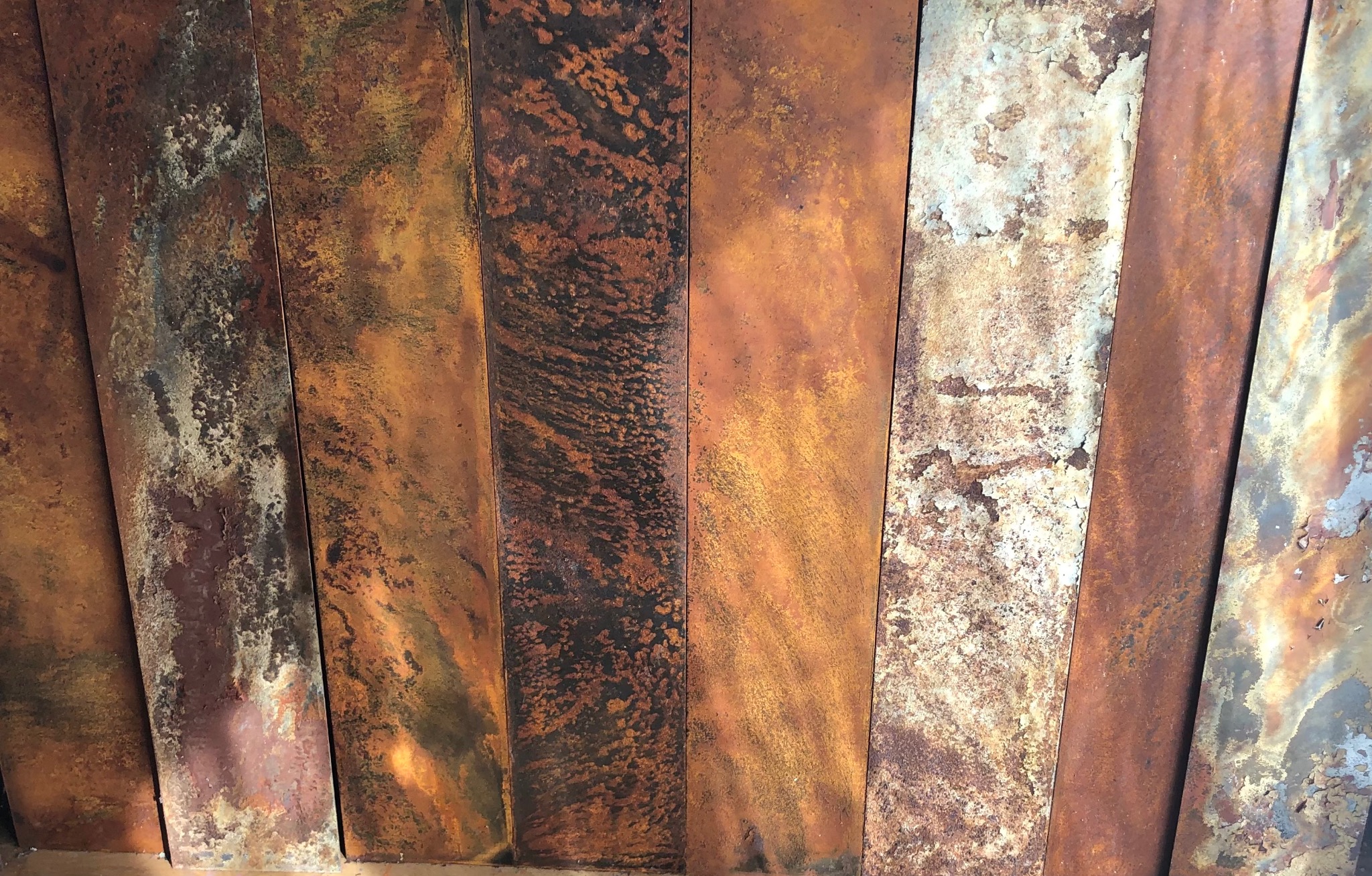 Rdzewiejące, pionowe metalowe panele z wyraźną teksturą i zróżnicowaną kolorystyką odcieni brązu, pomarańczy i szarości, ukazujące proces korozji