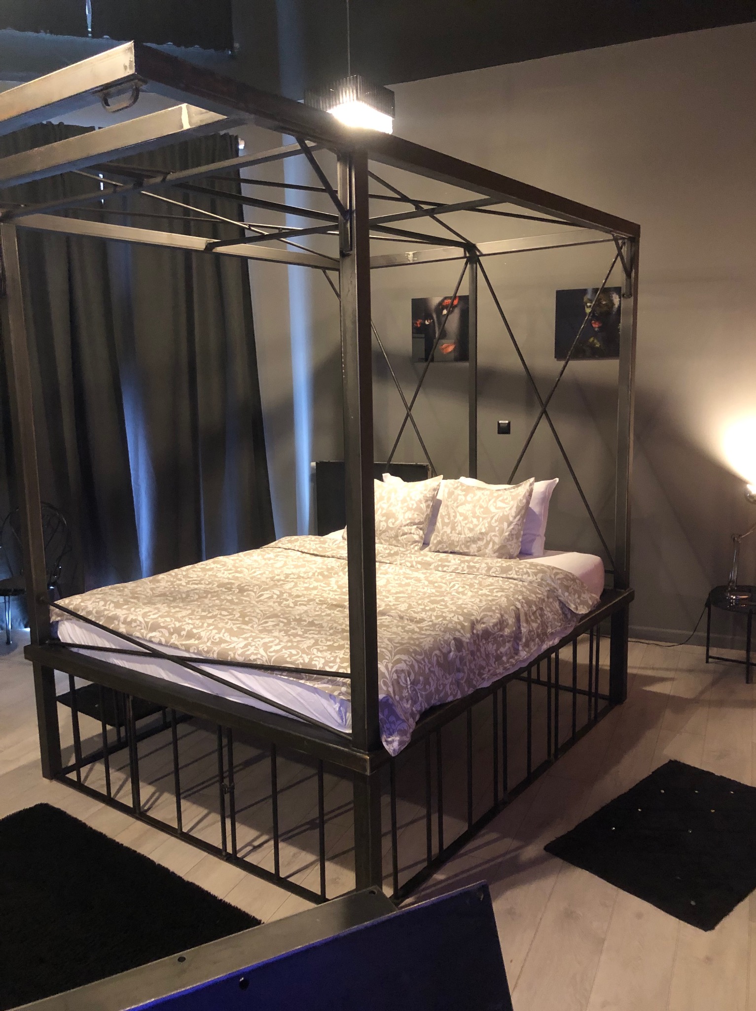 Metalowa rama łóżka z baldachimem, pomalowana na ciemny kolor, z pościelą w delikatny wzór, na podłodze czarne dywaniki, w tle szare ściany z obrazami.