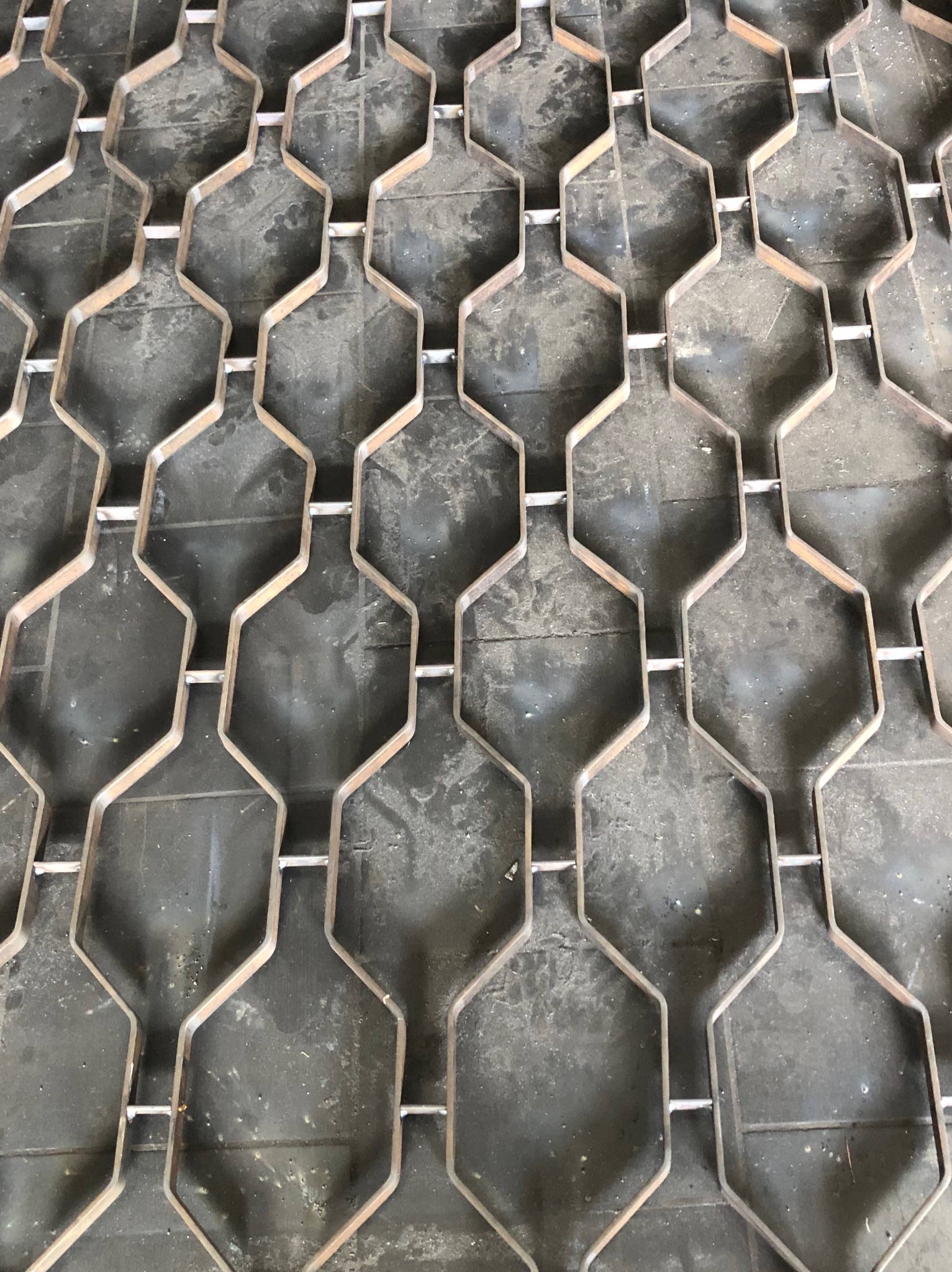 Metalowa konstrukcja o heksagonalnych oczkach, zgrzewana w punktach, leżąca na brudnej, ciemnej powierzchni.