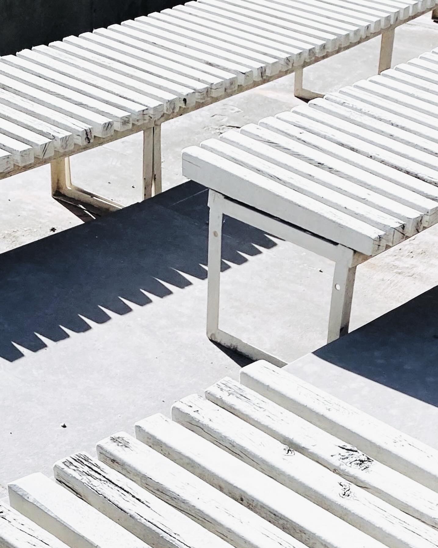 Białe, drewniane ławki parkowe w ostrym słońcu, rzucające wyraźne cienie na betonowe podłoże, z widoczną strukturą drewna i minimalistyczną konstrukcją.