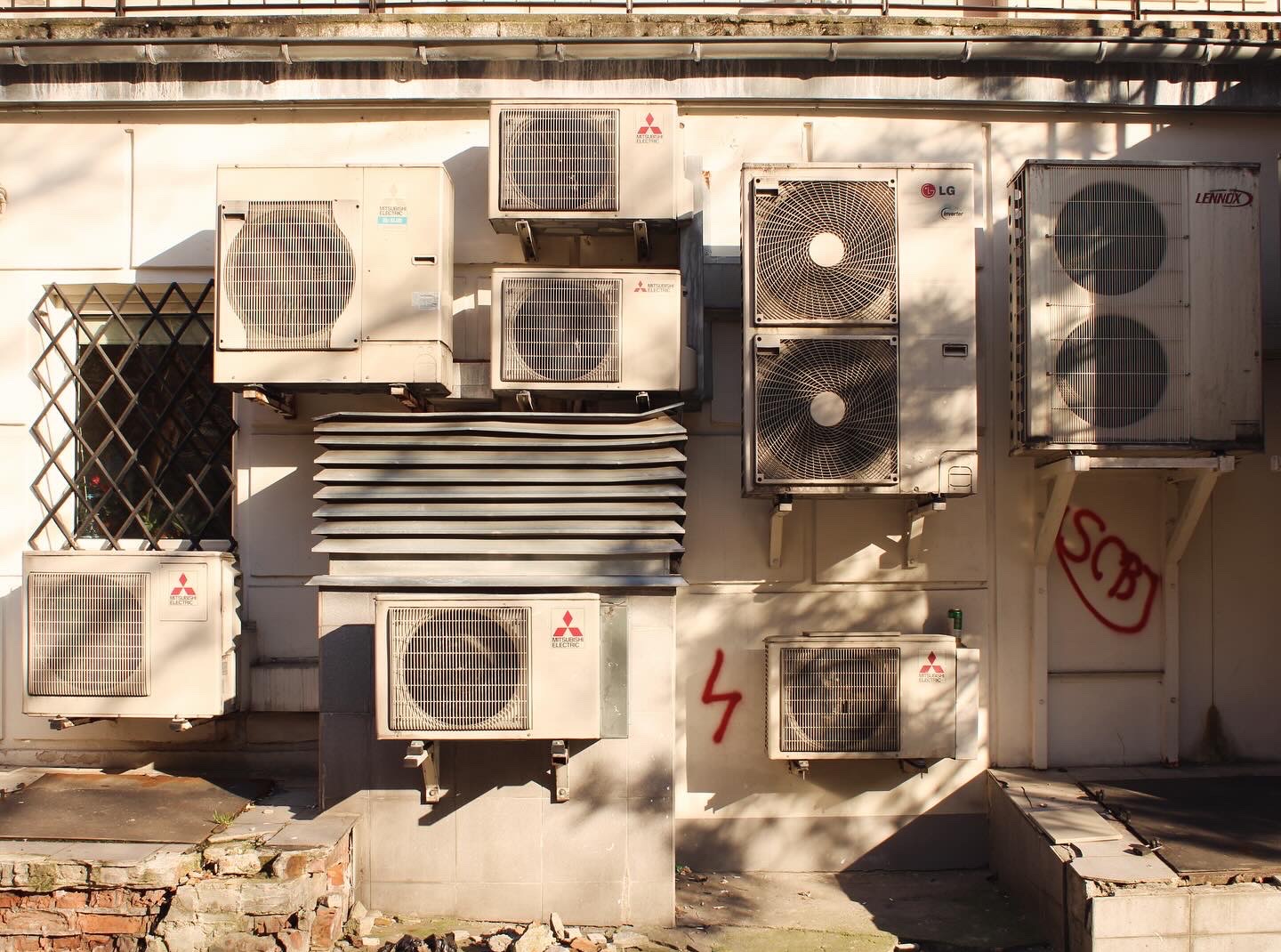 Ściana budynku z zamontowanymi różnymi modelami klimatyzatorów, w tym Mitsubishi Electric, LG i Lennox, z widocznymi śladami użytkowania i graffiti.