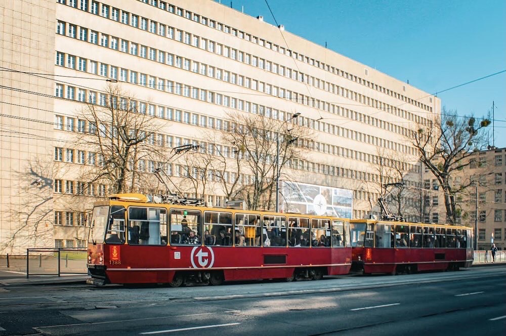 Czerwono-żółty tramwaj numer 1388 z pasażerami na tle jasnego budynku z wieloma oknami i drzewami bez liści.