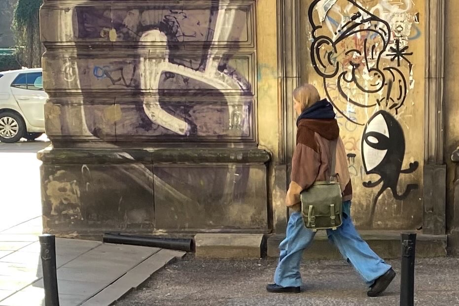 Osoba przechodzi obok ściany z graffiti przedstawiającego postacie z kreskówek i abstrakcyjne wzory. Widoczny samochód w tle.