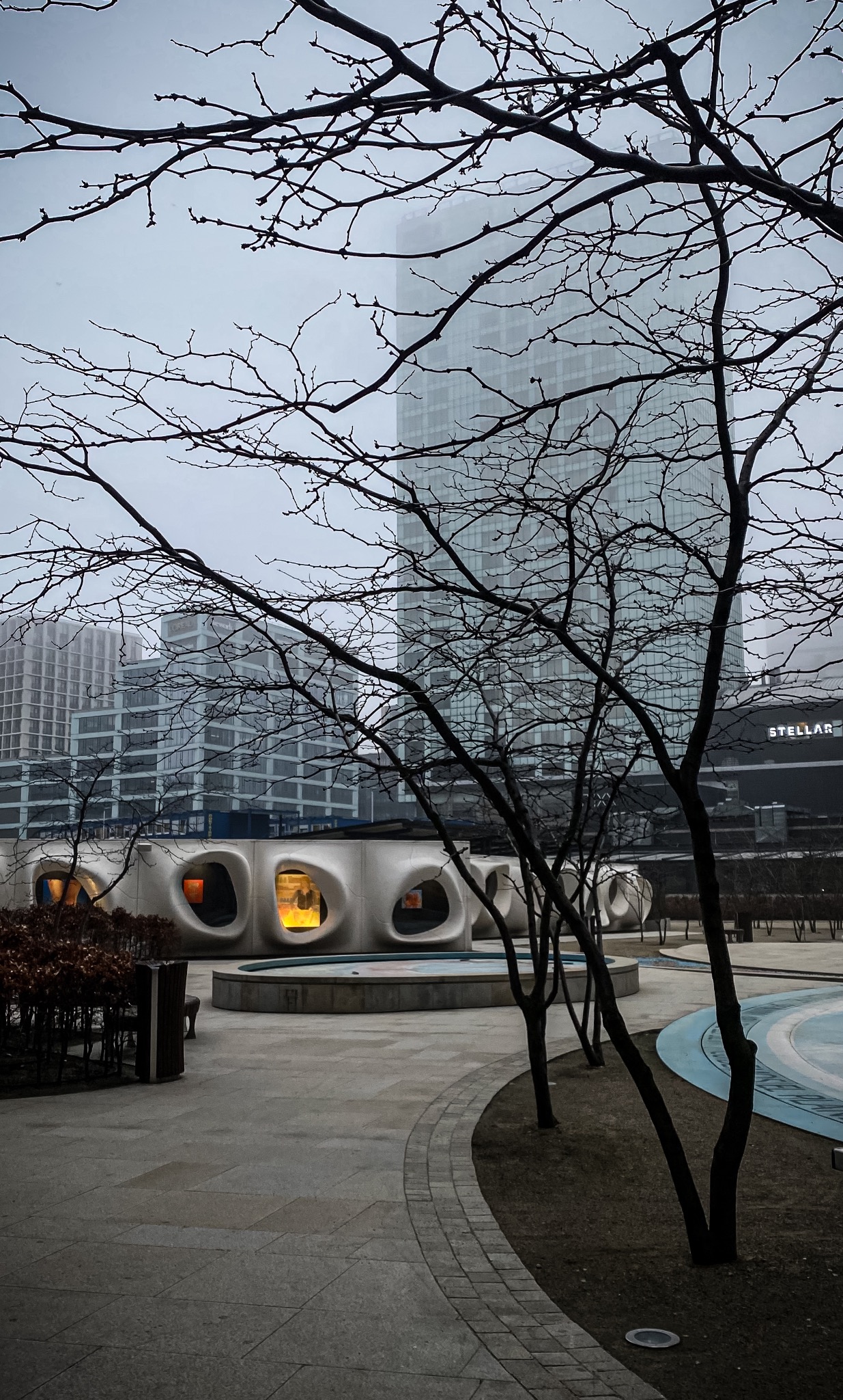 Nowoczesna architektura miejska w pochmurny dzień: biały budynek z okrągłymi otworami, wysokie biurowce w tle, nagie drzewa na pierwszym planie.