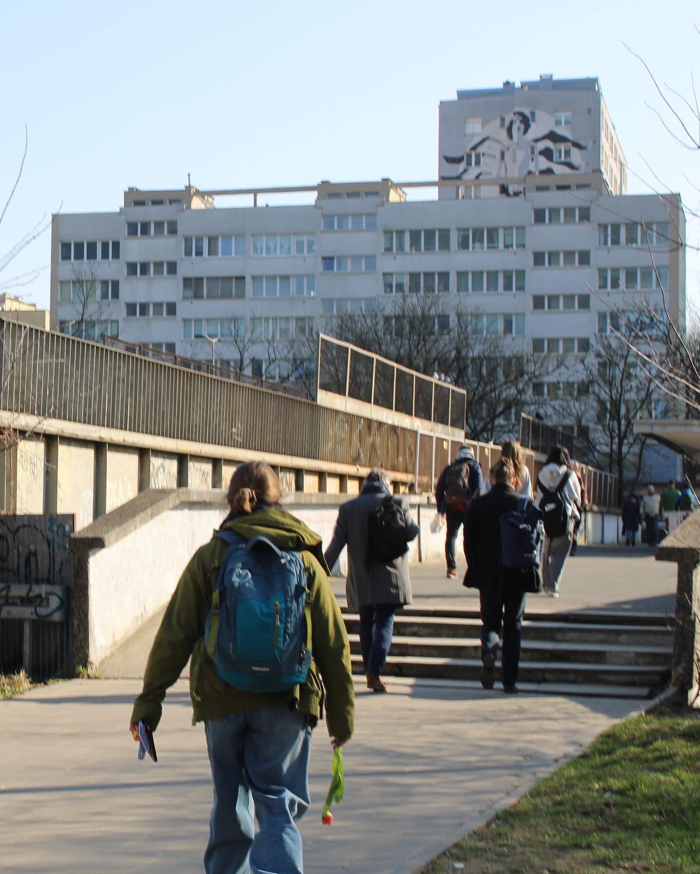 Grupa osób z plecakami wchodzi po schodach na wiadukt; w tle budynek z muralem.