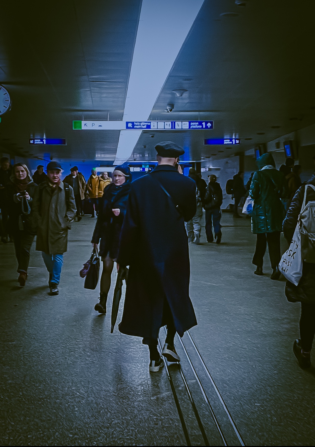 Tłum ludzi przechodzących przez podziemne przejście, widoczne znaki informacyjne i oświetlenie sufitowe.