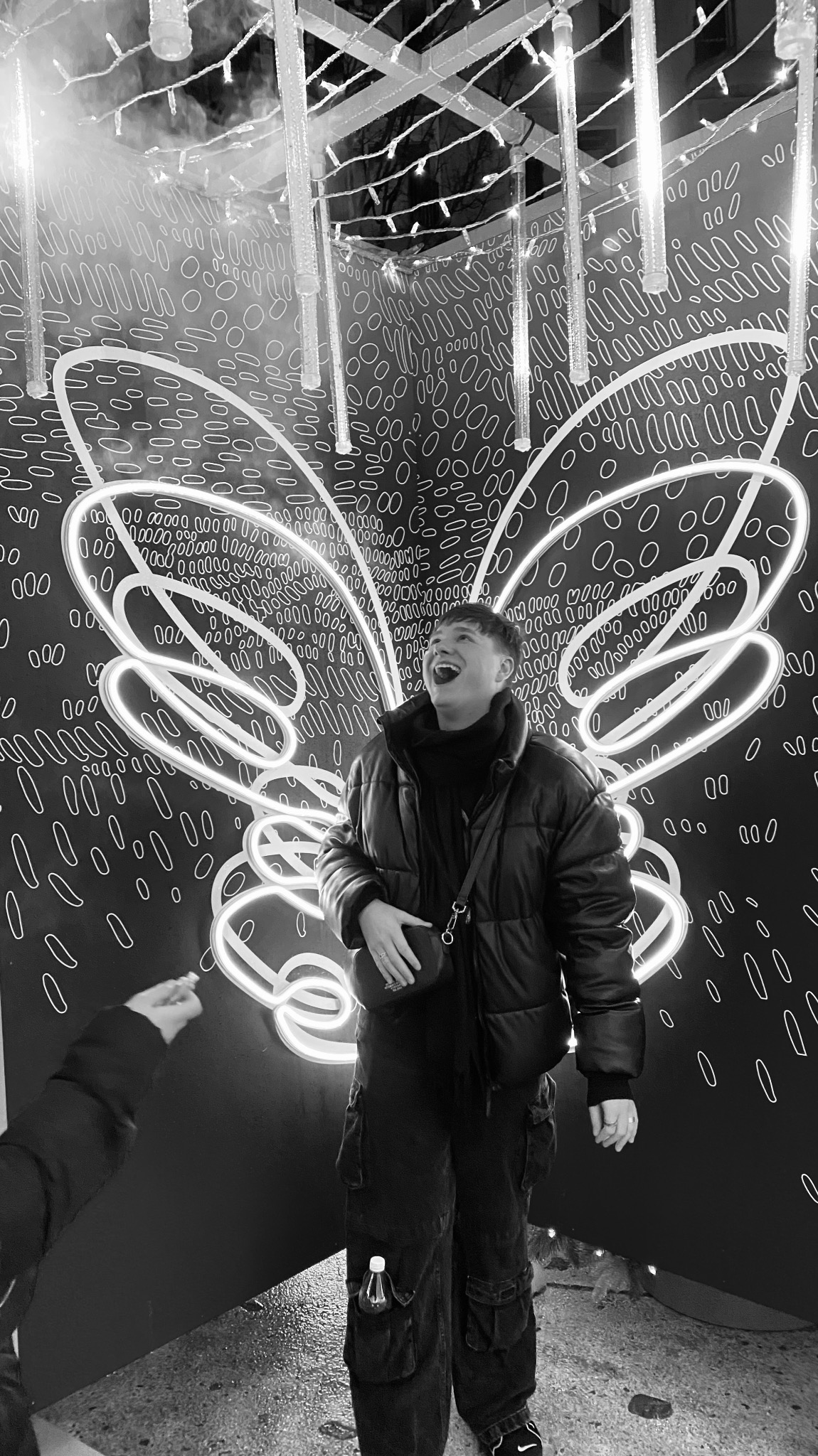 Osoba w kurtce śmiejąca się na tle neonowych skrzydeł motyla i abstrakcyjnego wzoru na ścianie podczas eventu.