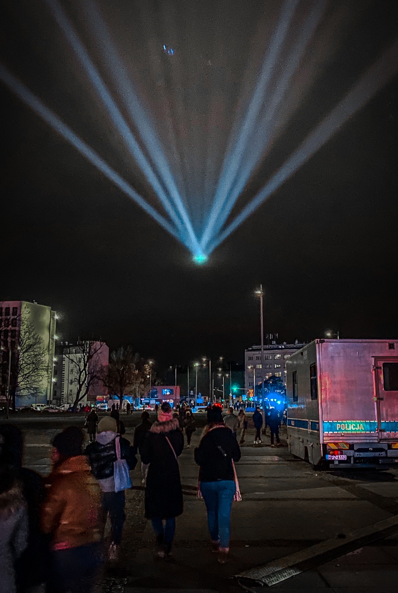 Nocne ujęcie miejskiej przestrzeni z tłumem ludzi, nad którą rozpościerają się promienie reflektorów, z radiowozem policyjnym w tle.