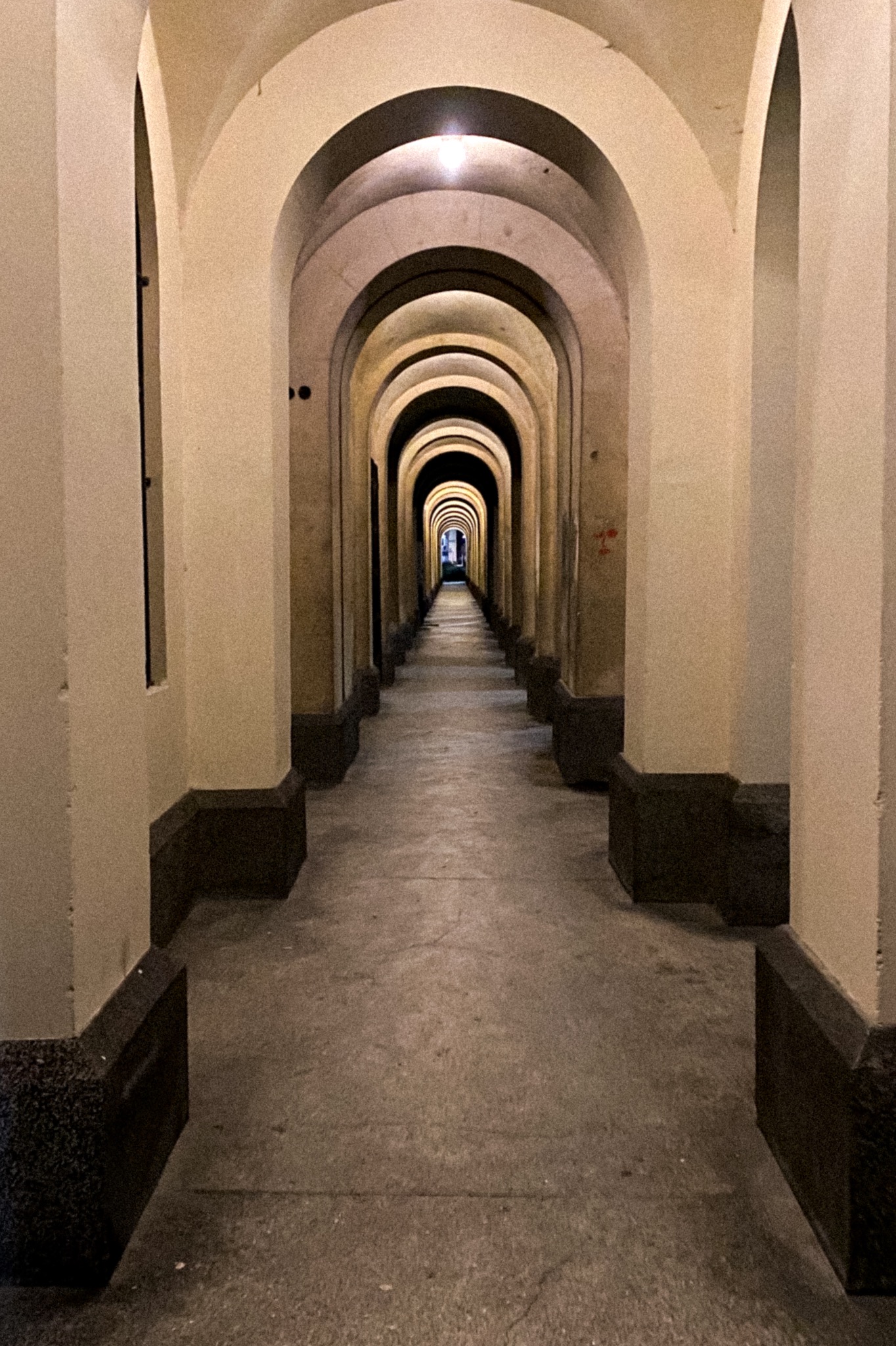 Długi korytarz z rzędem identycznych łuków, perspektywa zbieżna do punktu świetlnego na końcu.