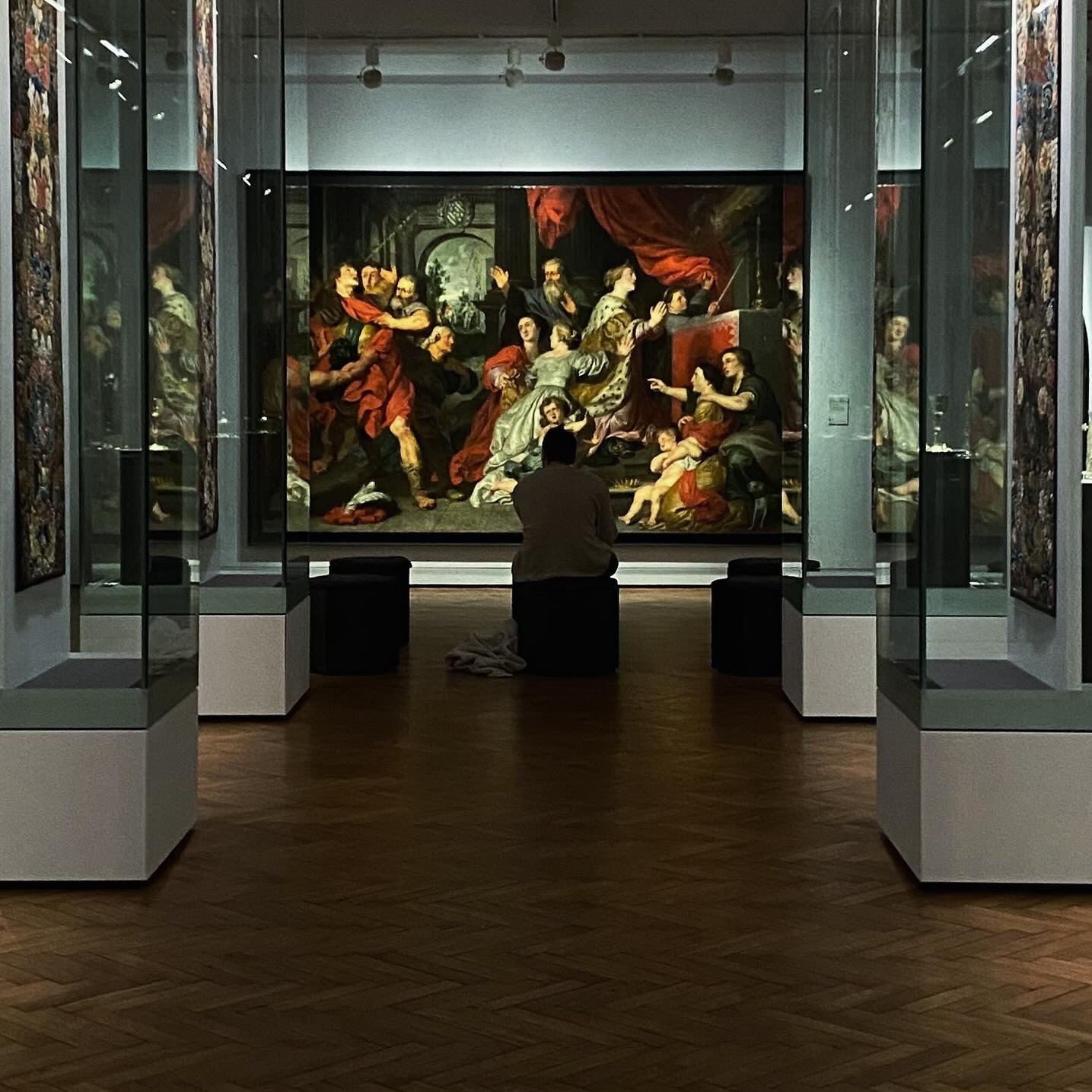 Osoba siedząca przed dużym obrazem w muzeum, otoczonym przez szklane gabloty, na parkiecie z ciemnego drewna.