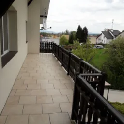 Beżowe płytki na balkonie z drewnianą balustradą i widokiem na okolicę, antena satelitarna na ścianie budynku.