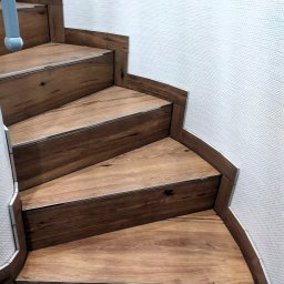 Usługi Remontowo-Wykończeniowe Paweł Czyrnek - Drewnopodobne płytki ułożone na schodach spiralnych z metalowymi listwami krawędziowymi, przylegające do teksturowanej ściany.