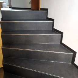 Usługi Remontowo-Wykończeniowe Paweł Czyrnek - Schody wyłożone ciemnoszarą płytką z metalowymi listwami na krawędziach stopni, widok z dołu, fragment ściany z białym tynkiem i fragment drzwi.