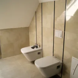 Jasna łazienka na poddaszu ze skośnym sufitem, z beżowymi płytkami ułożonymi w dwóch wzorach: duże płyty i mozaika oddzielone czarnymi listwami. Wisząca toaleta i bidet z chromowaną baterią.