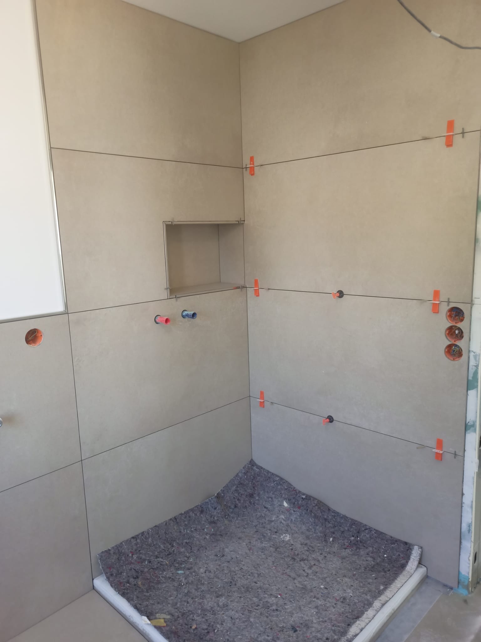 Ściany łazienki w trakcie układania beżowych płytek wielkoformatowych, widoczne krzyżyki dystansowe, poziomica laserowa, wnęka w ścianie przygotowana pod półkę, zabezpieczona podłoga.