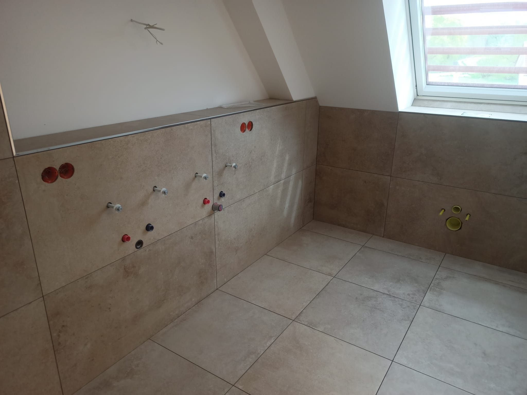 Łazienka w trakcie remontu w poddaszu: beżowe płytki na ścianach i podłodze, widoczne przyłącza wodne i kanalizacyjne, okno dachowe z żaluzjami.