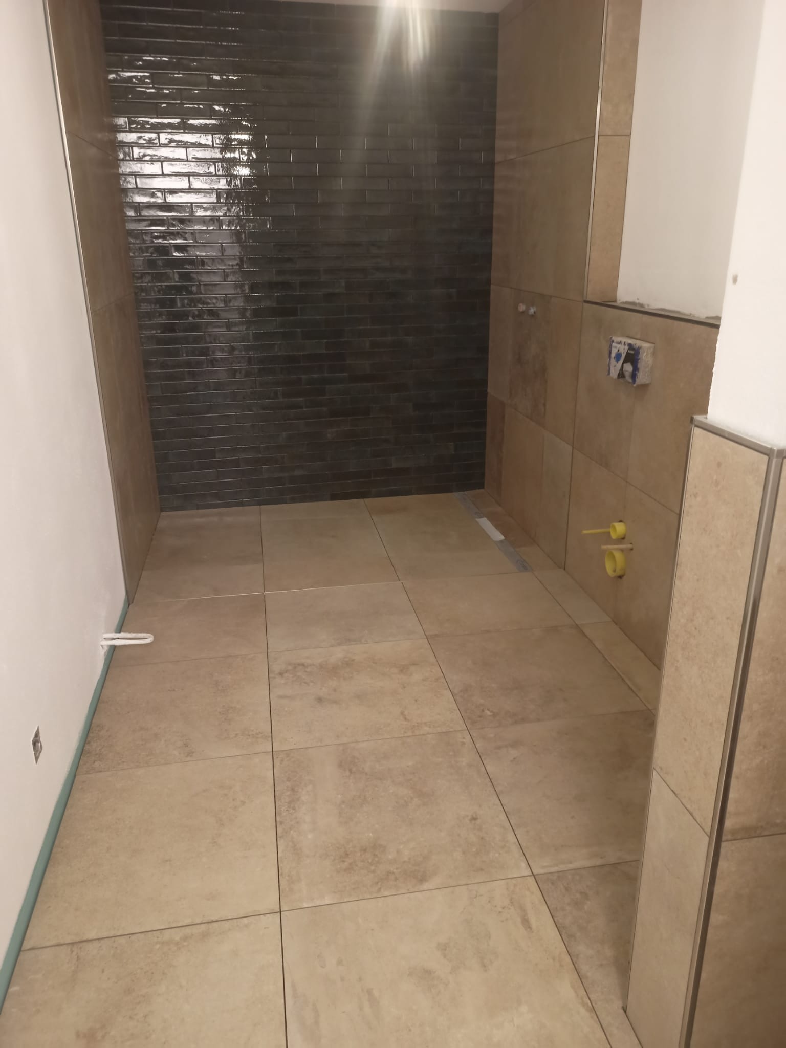 Wnętrze łazienki w trakcie prac wykończeniowych, widoczne ciemne, błyszczące płytki na ścianie kabiny prysznicowej oraz beżowe płytki podłogowe i ścienne. Na ścianie wystają żółte końcówki rur...