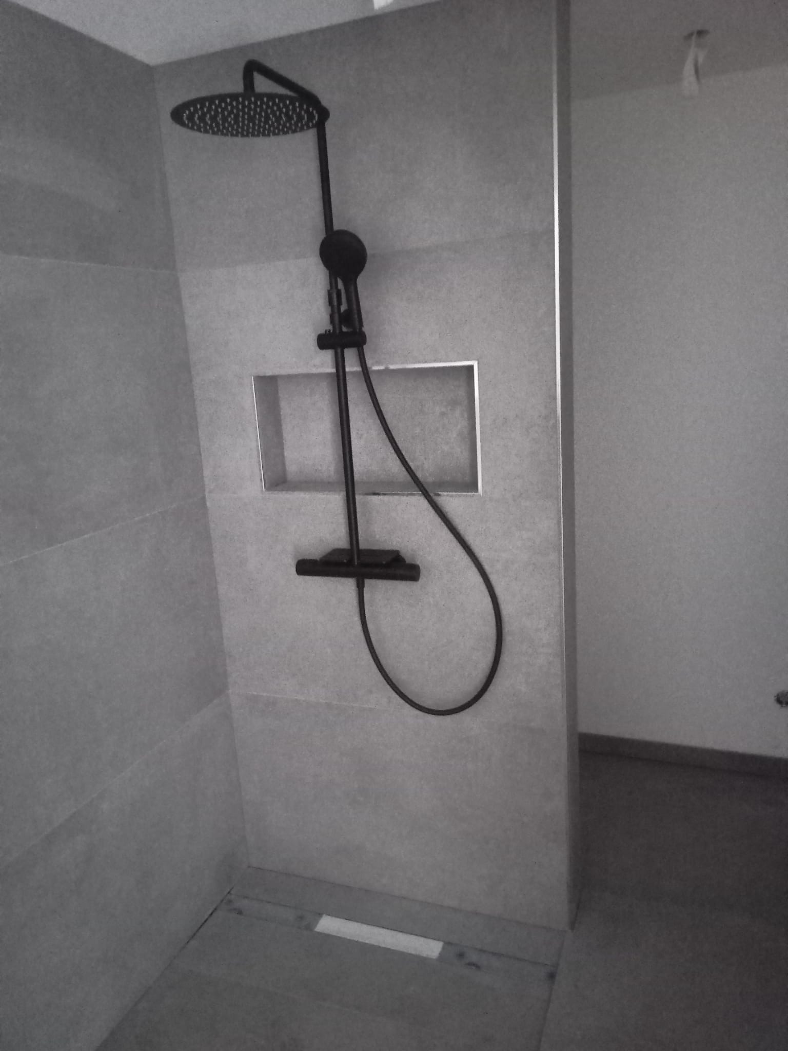 Nowoczesny prysznic z czarną armaturą i deszczownicą w łazience wykończonej szarymi płytkami, z wnęką w ścianie i odpływem liniowym w podłodze.