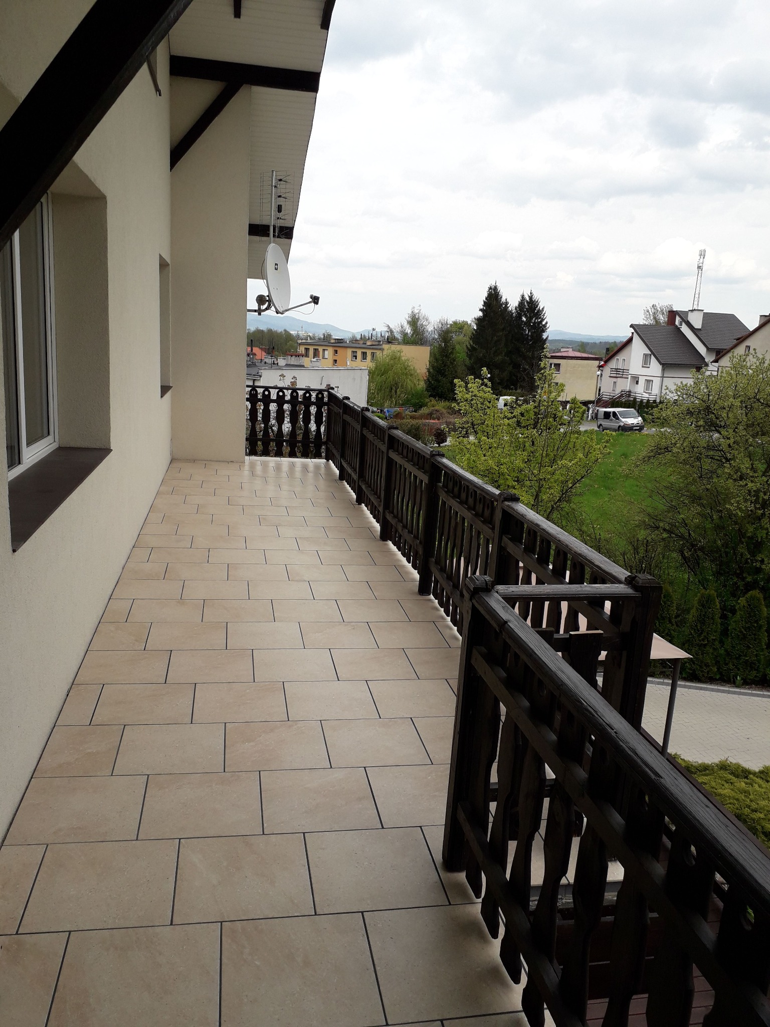 Beżowe płytki na balkonie z drewnianą balustradą i widokiem na okolicę, antena satelitarna na ścianie budynku.