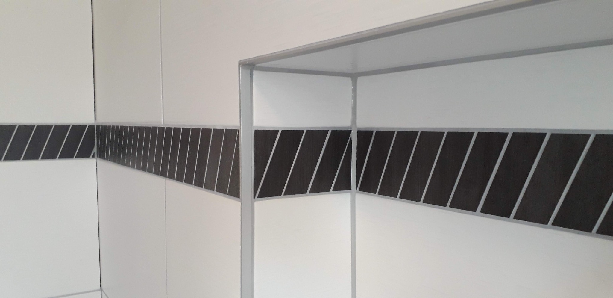 Białe płytki ścienne z czarnym pasem dekoracyjnym z ukośnymi paskami, narożna półka z płytek w łazience.