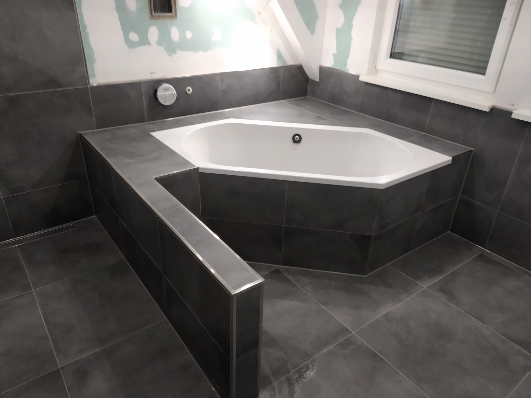 Narożna wanna obudowana ciemnymi płytkami w łazience w trakcie wykańczania; widoczne niedokończone ściany i biała stolarka okienna.