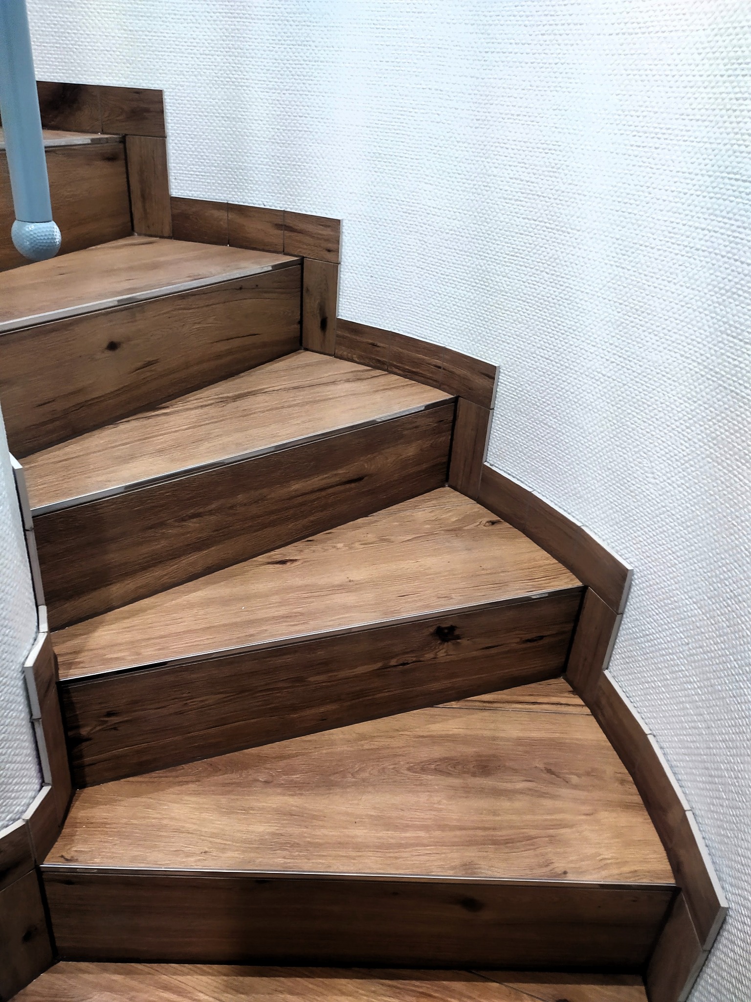 Drewnopodobne płytki ułożone na schodach spiralnych z metalowymi listwami krawędziowymi, przylegające do teksturowanej ściany.