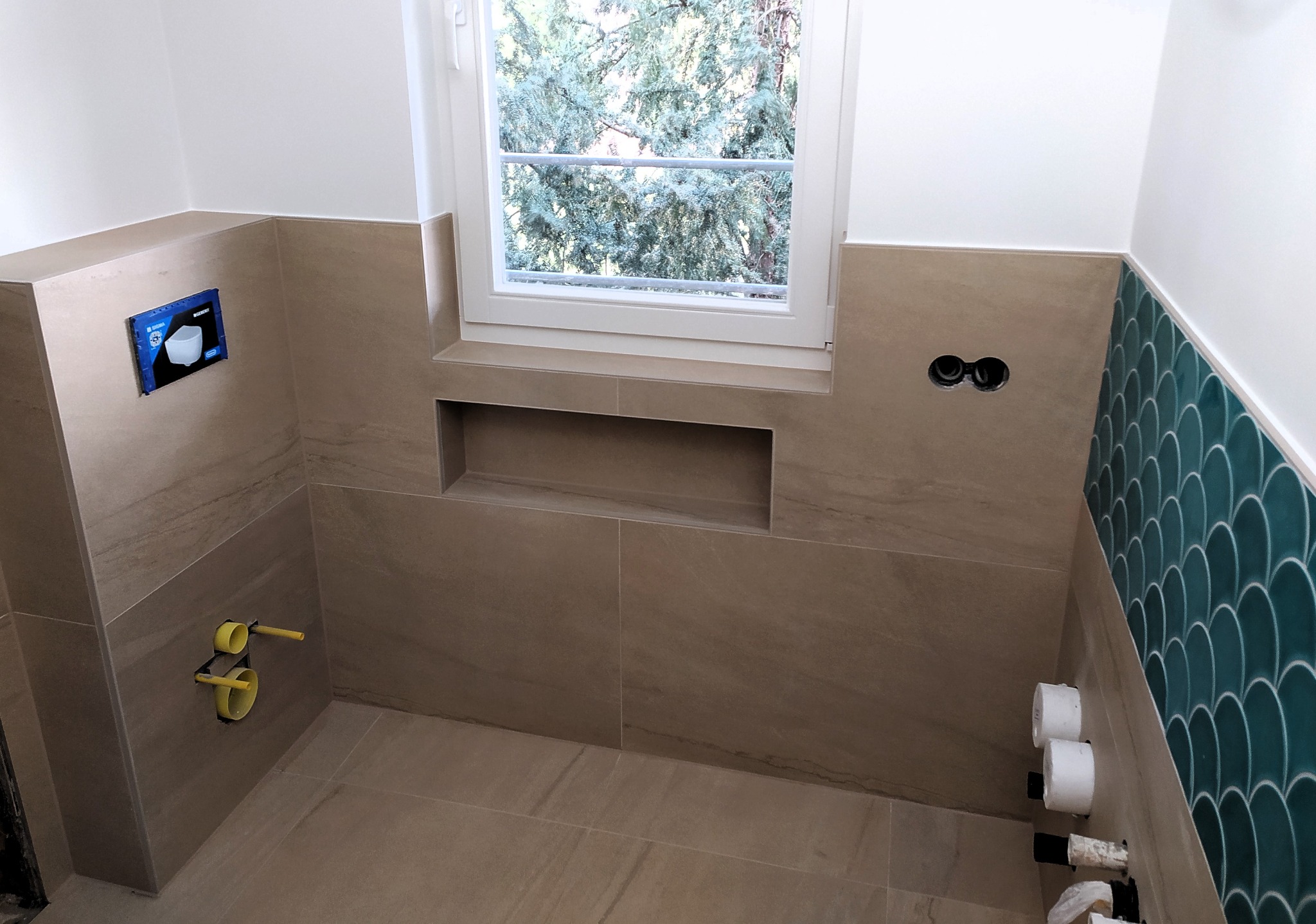 Wnętrze łazienki w trakcie wykańczania: beżowe płytki imitujące kamień na ścianach i podłodze, fragment ściany wyłożony turkusową mozaiką w kształcie rybiej łuski, okno z widokiem na drzewo...