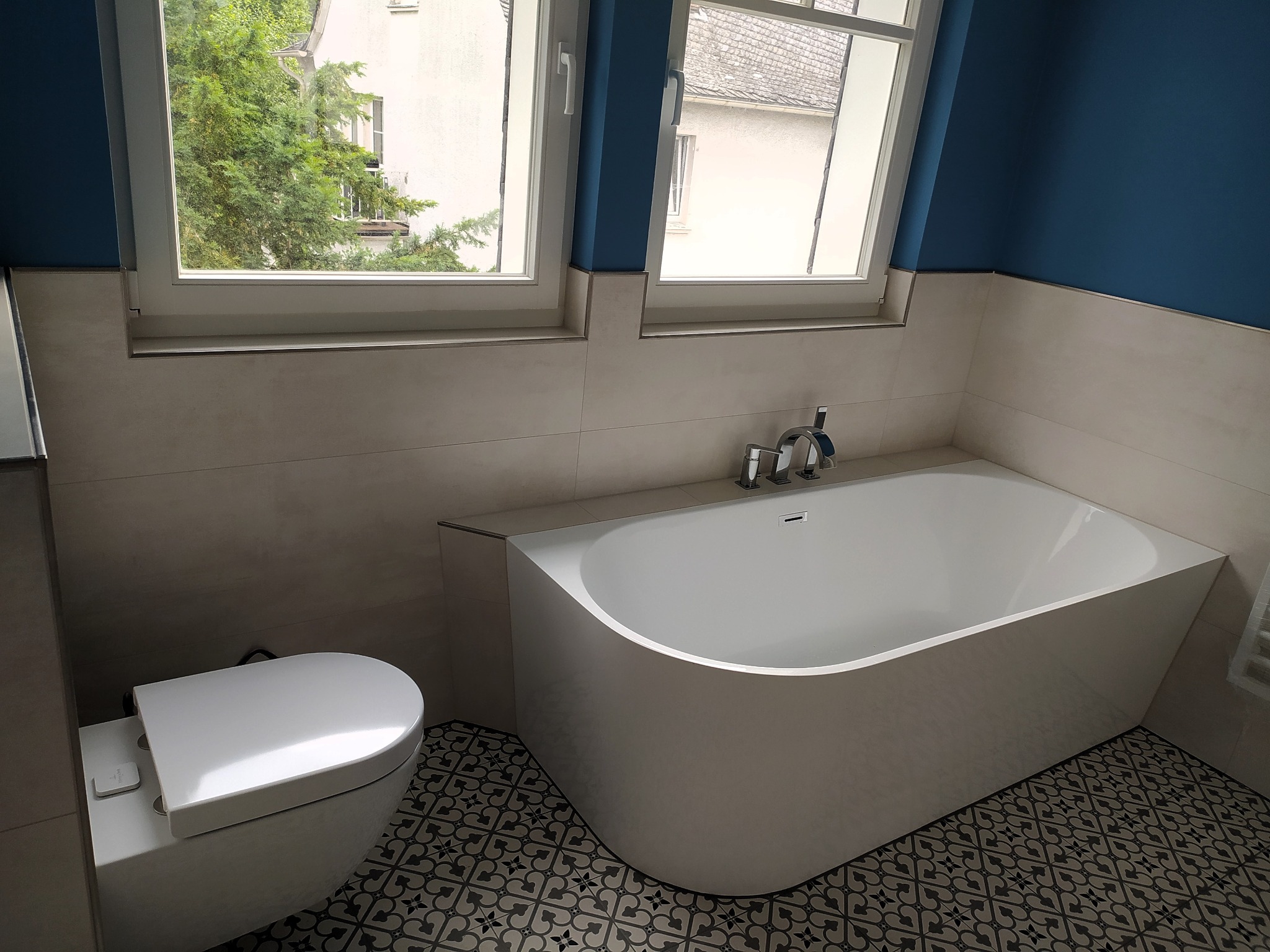 Nowoczesna łazienka z wolnostojącą wanną, wiszącą toaletą i wzorzystą podłogą, okno z widokiem na zieleń