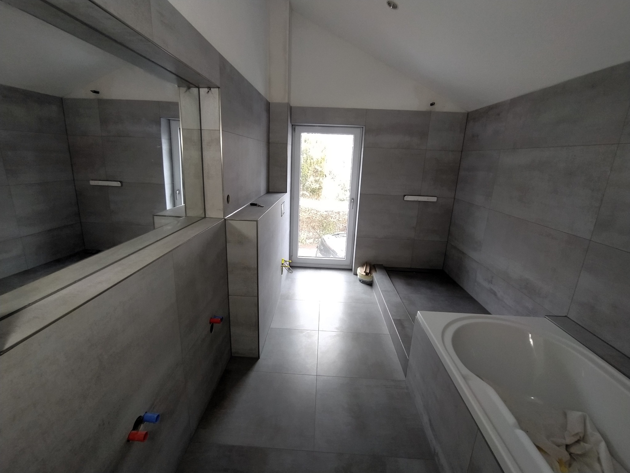 Szara łazienka z dużymi płytkami, widoczne podejścia wodne z czerwono-niebieskimi zaworami, biała wanna i okno z widokiem na zewnątrz, częściowo wykończona.