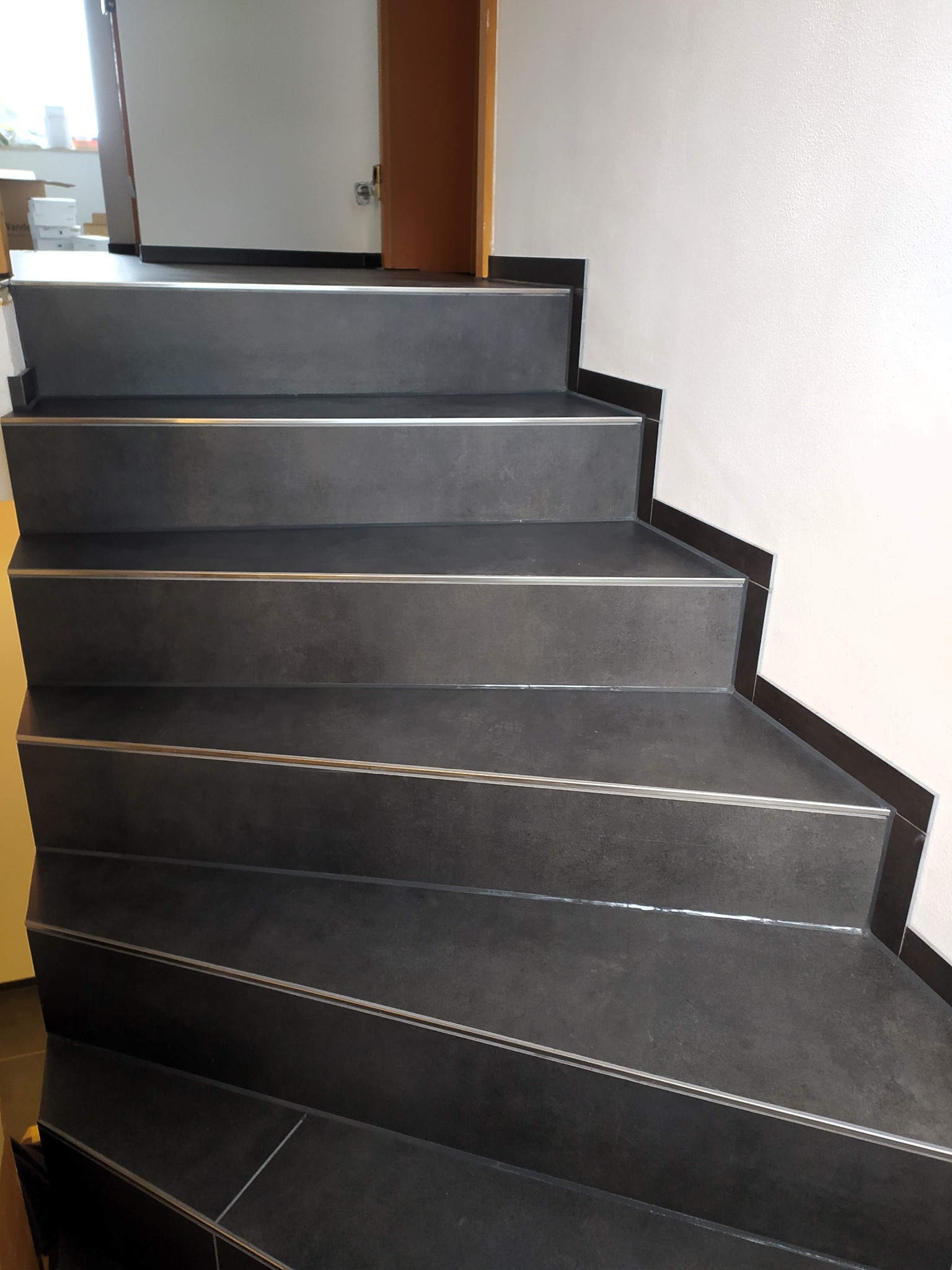 Schody wyłożone ciemnoszarą płytką z metalowymi listwami na krawędziach stopni, widok z dołu, fragment ściany z białym tynkiem i fragment drzwi.