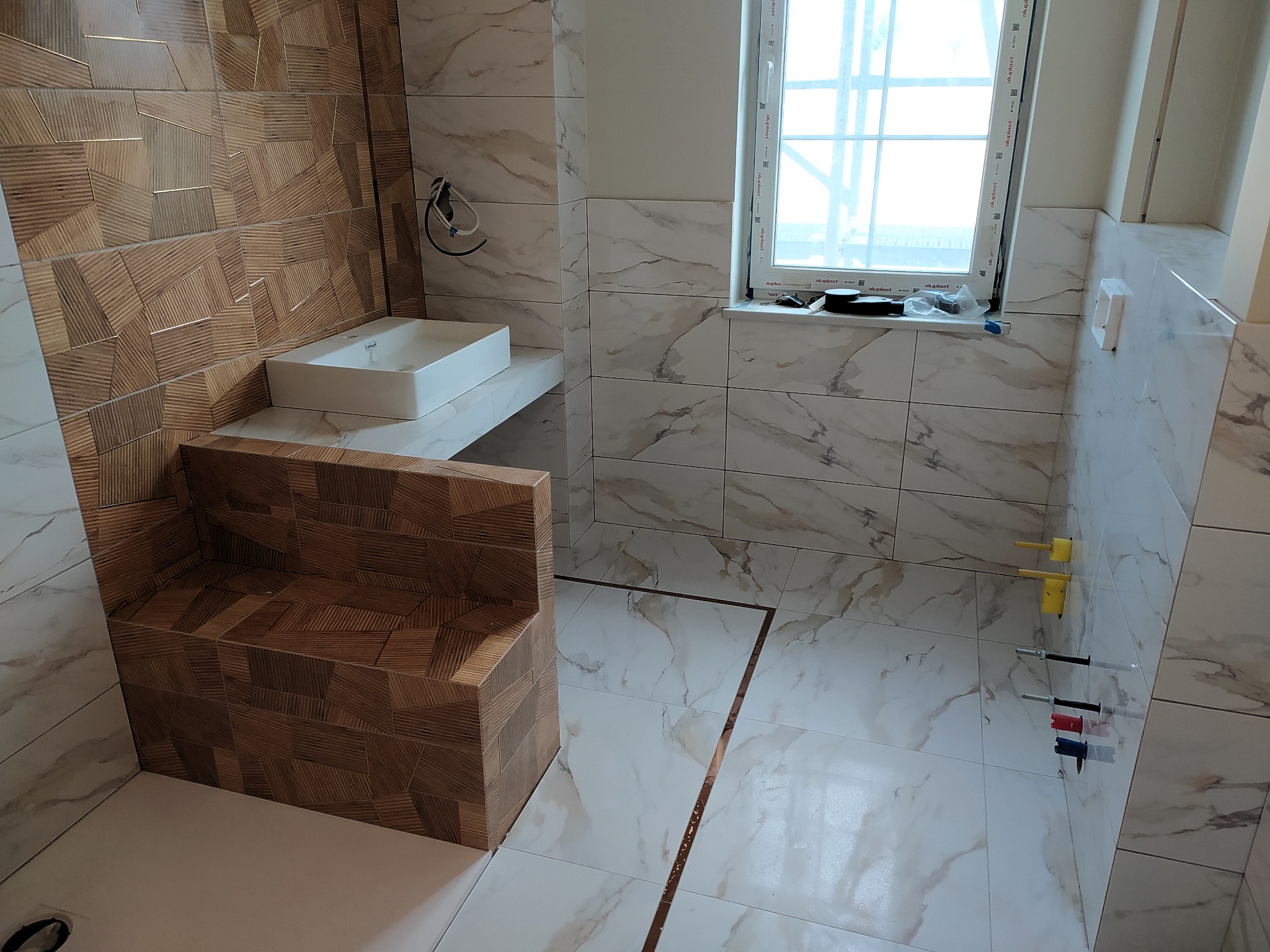 Nowoczesna łazienka w trakcie wykańczania: białe marmurowe płytki, drewnopodobna mozaika na ścianie i obudowie wanny, biała umywalka na blacie, okno z widokiem na zewnątrz, widoczne przyłącza wodne.