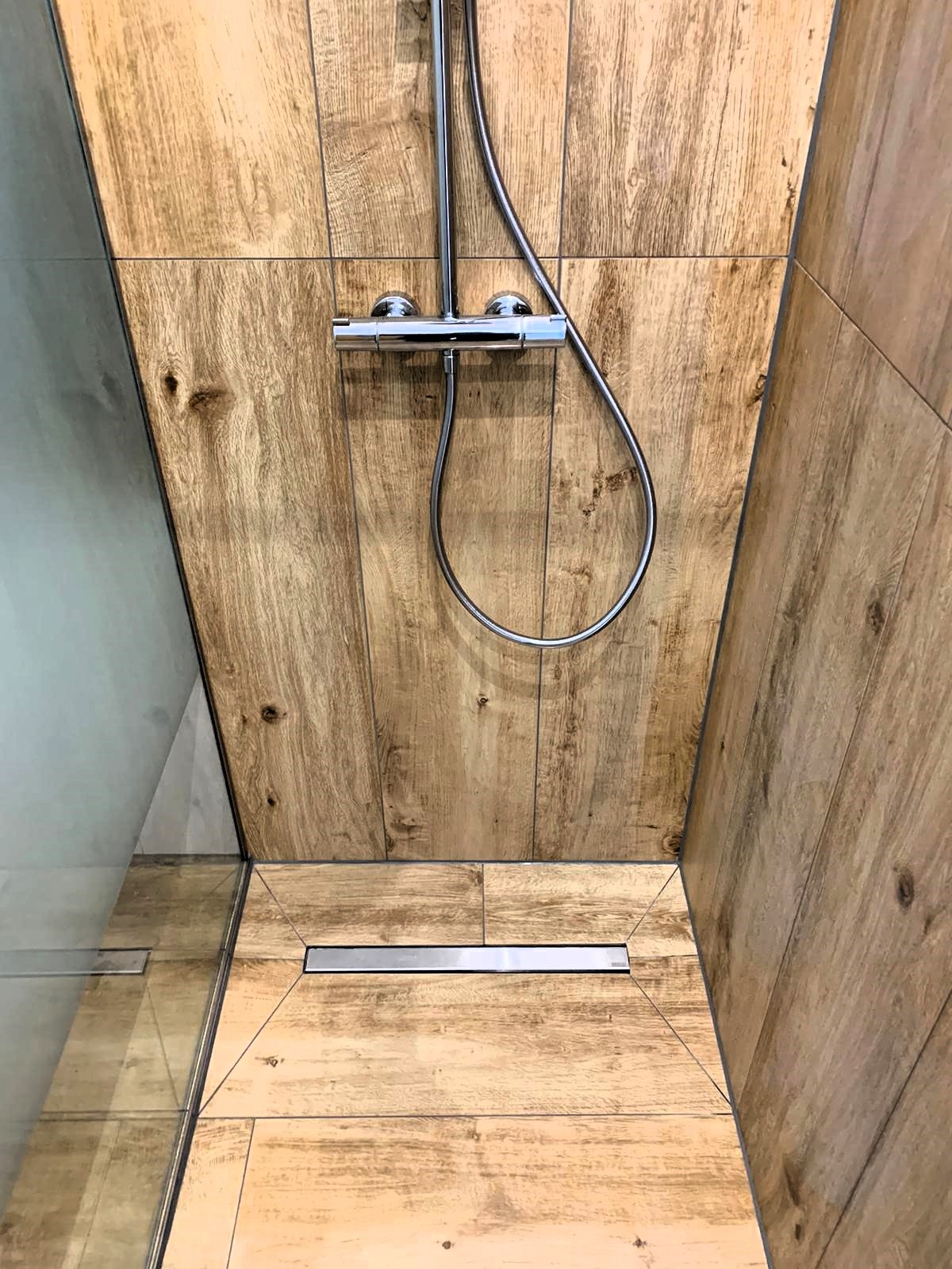 Kabina prysznicowa wyłożona płytkami imitującymi drewno, z chromowaną armaturą i odpływem liniowym w podłodze.