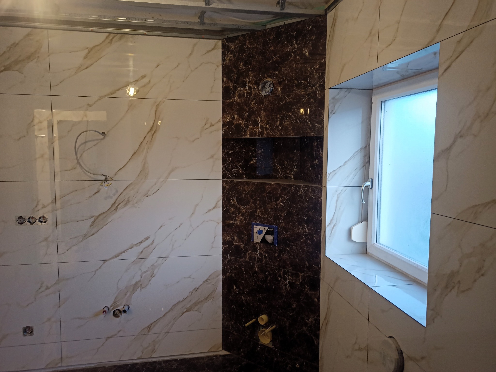 Łazienka w trakcie wykańczania: ściany wyłożone płytkami imitującymi marmur w odcieniach bieli i brązu, wnęka z ciemnobrązowymi płytkami, przygotowane instalacje elektryczne i hydrauliczne, okno...