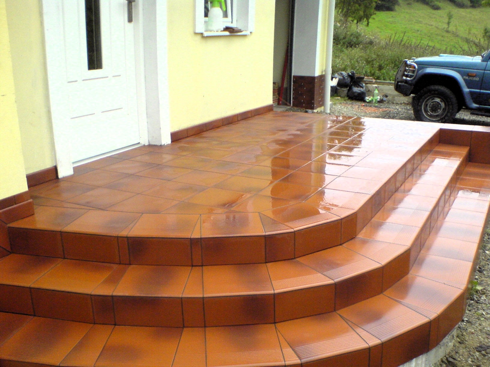 Błyszczący, brązowy taras z płytek ceramicznych z trzema schodkami prowadzącymi do drzwi wejściowych, widoczne odbicia na mokrej powierzchni, w tle zaparkowany samochód i worki ze śmieciami.