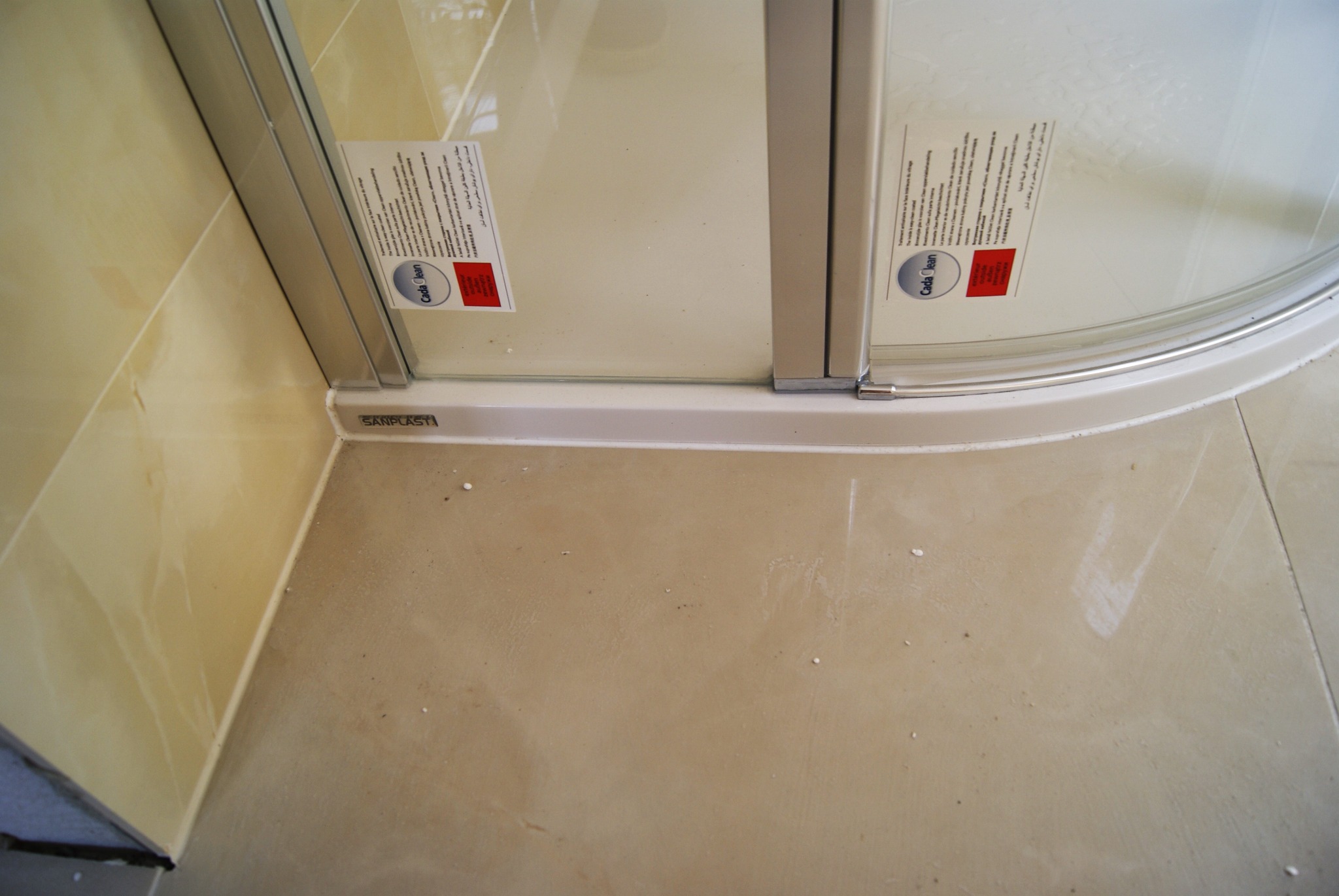 Fragment narożnika kabiny prysznicowej Sanplast z naklejkami informacyjnymi Cada Clean, widoczne łączenie płytek podłogowych i ściennych w odcieniach beżu.