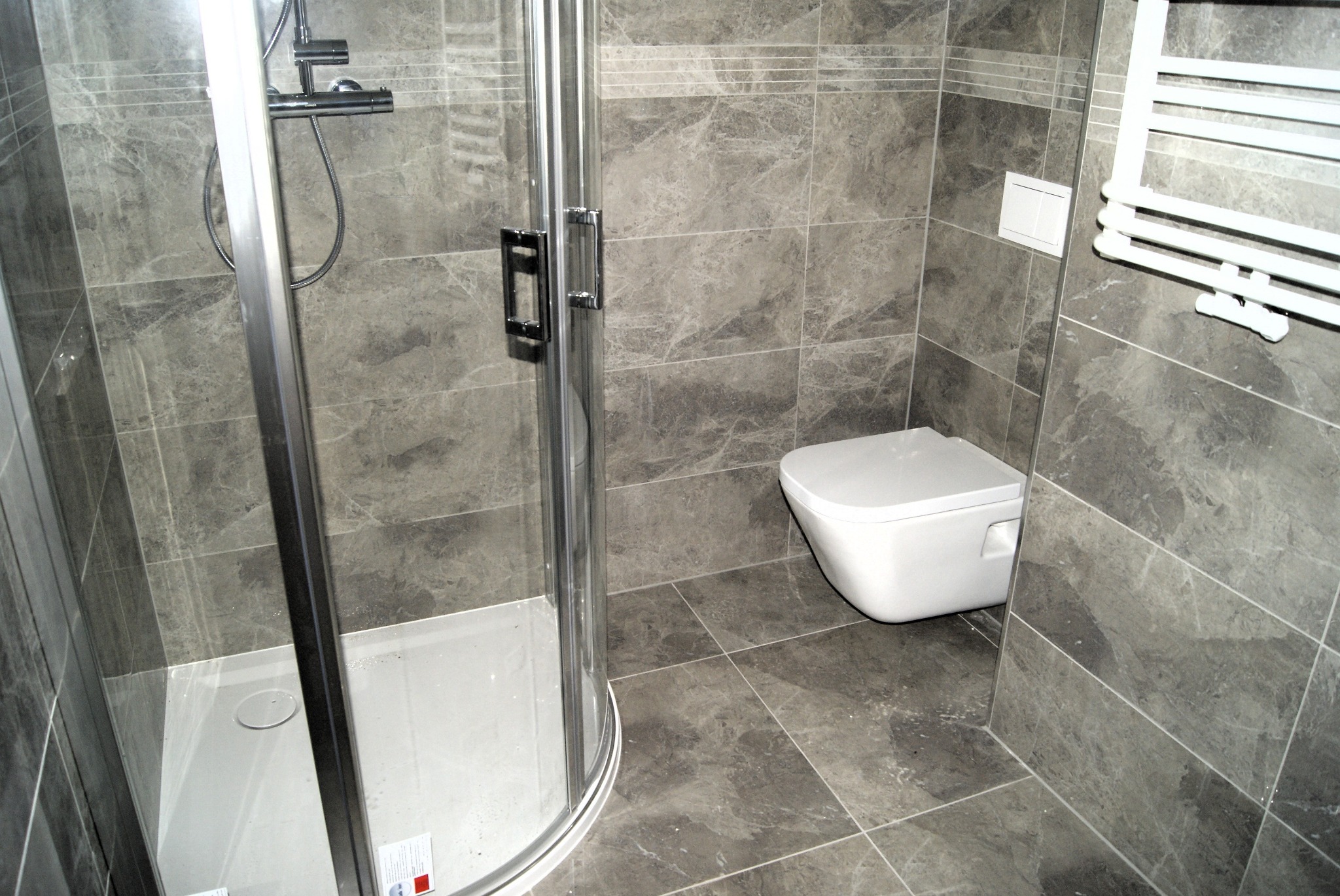 Szara łazienka z kabiną prysznicową z chromowanymi profilami, podwieszaną toaletą i grzejnikiem drabinkowym, wykończona płytkami imitującymi marmur z delikatną fugą.