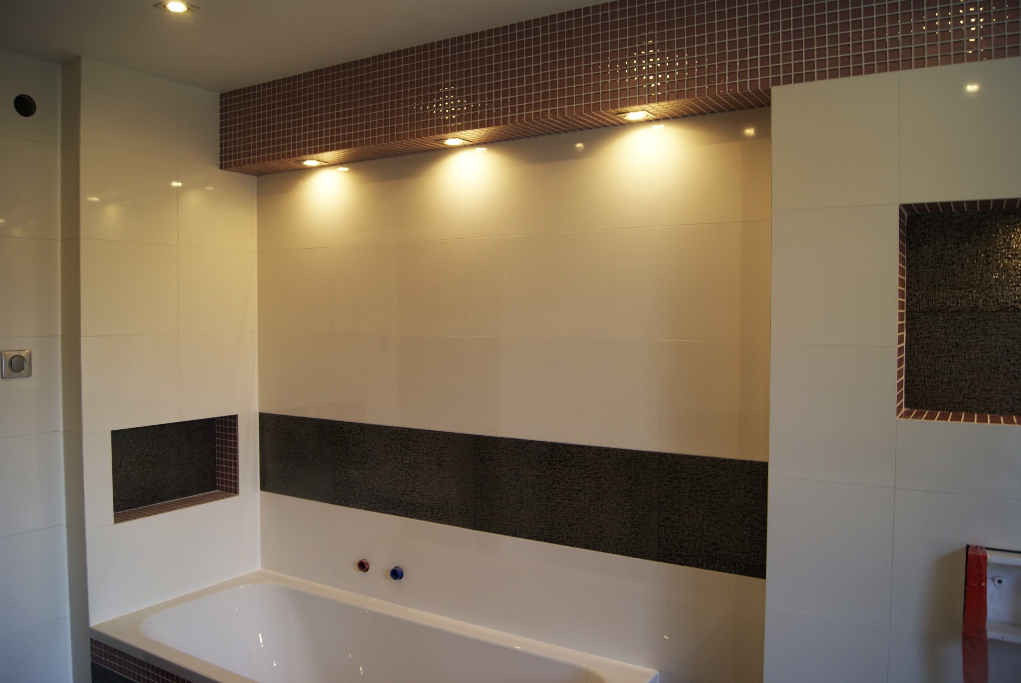 Wykończona łazienka z biało-beżowymi płytkami i mozaiką w odcieniach brązu, podświetlona górnymi halogenami, z wnęką wykończoną mozaiką i widoczną częścią białej wanny.