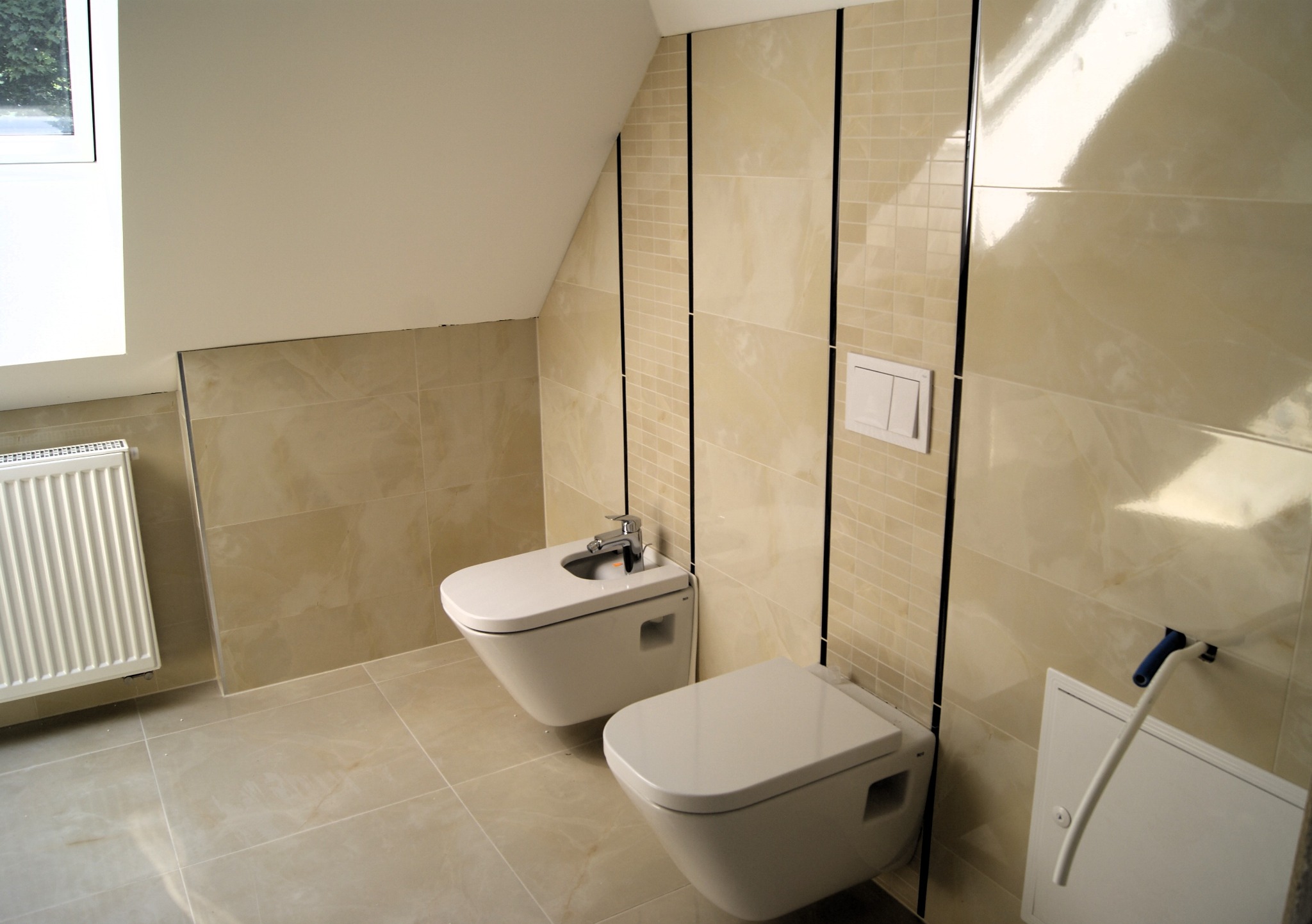 Jasna łazienka na poddaszu ze skośnym sufitem, z beżowymi płytkami ułożonymi w dwóch wzorach: duże płyty i mozaika oddzielone czarnymi listwami. Wisząca toaleta i bidet z chromowaną baterią.